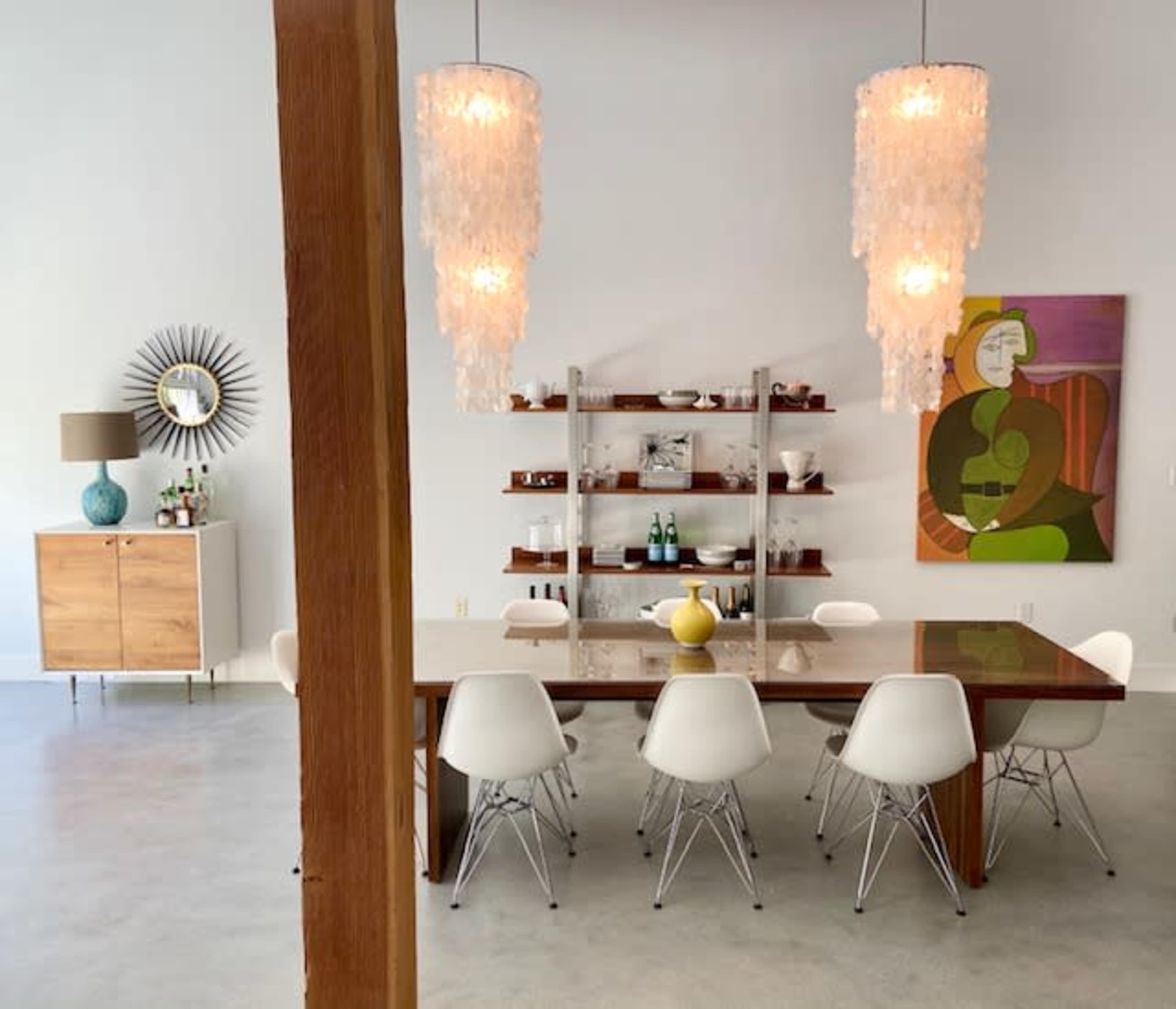 A modern dining area features a wooden table surrounded by white chairs, with two large pendant lights and a colorful wall painting in the background.