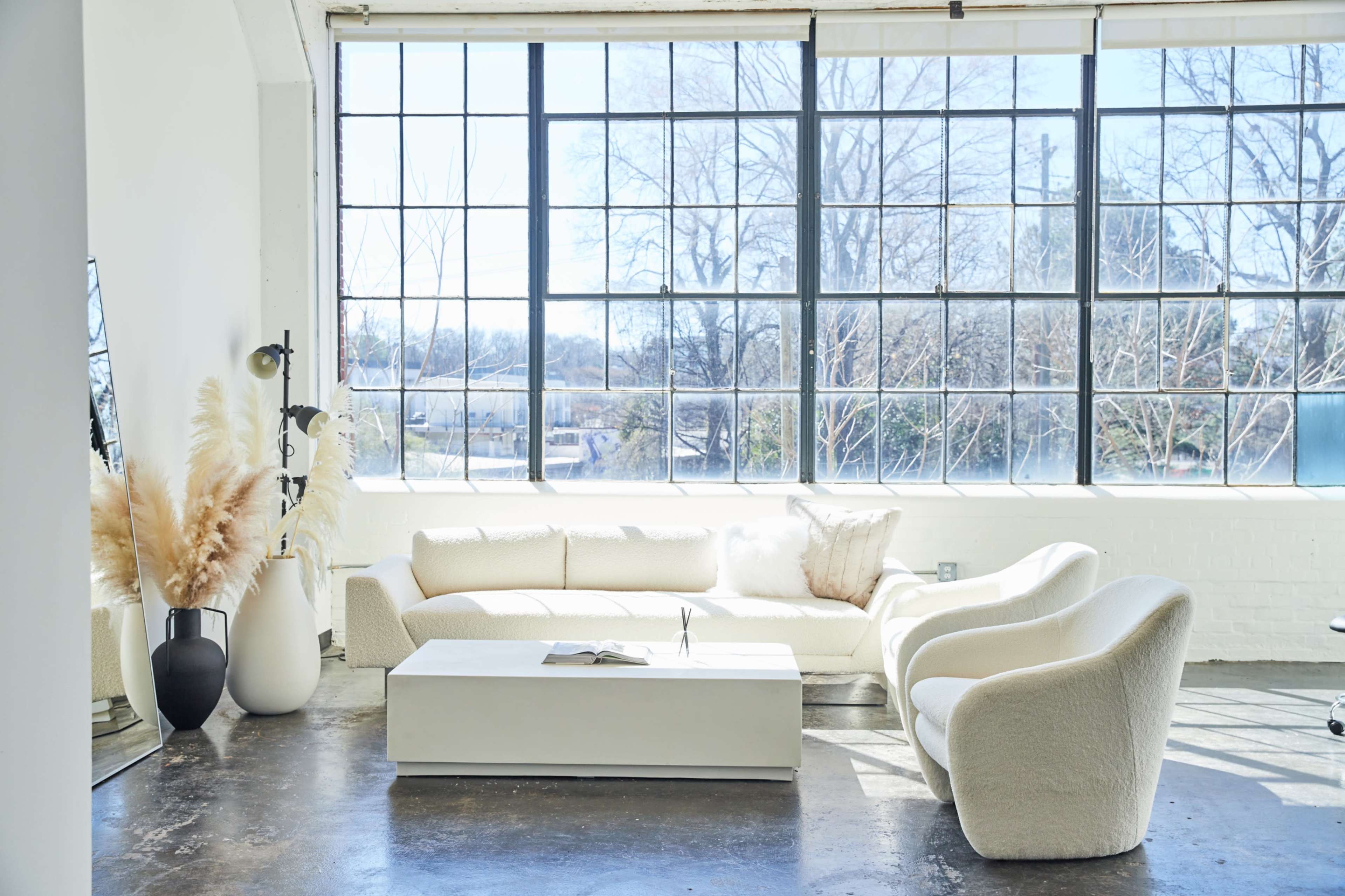 A bright living room features a white, plush sofa, a minimalist coffee table, and large windows allowing natural light to fill the space.