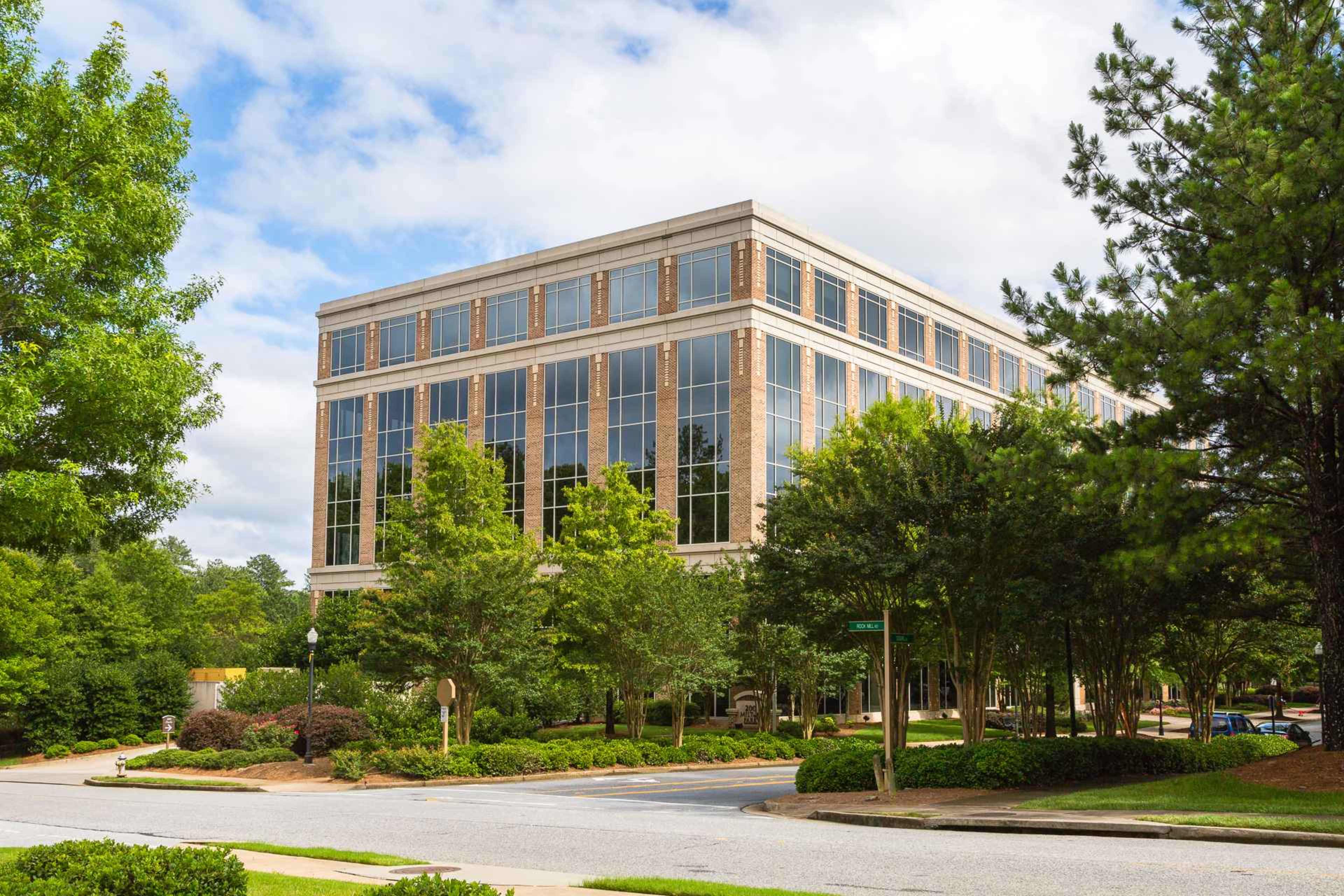 A modern office building with large windows surrounded by trees and parking areas near a street intersection.