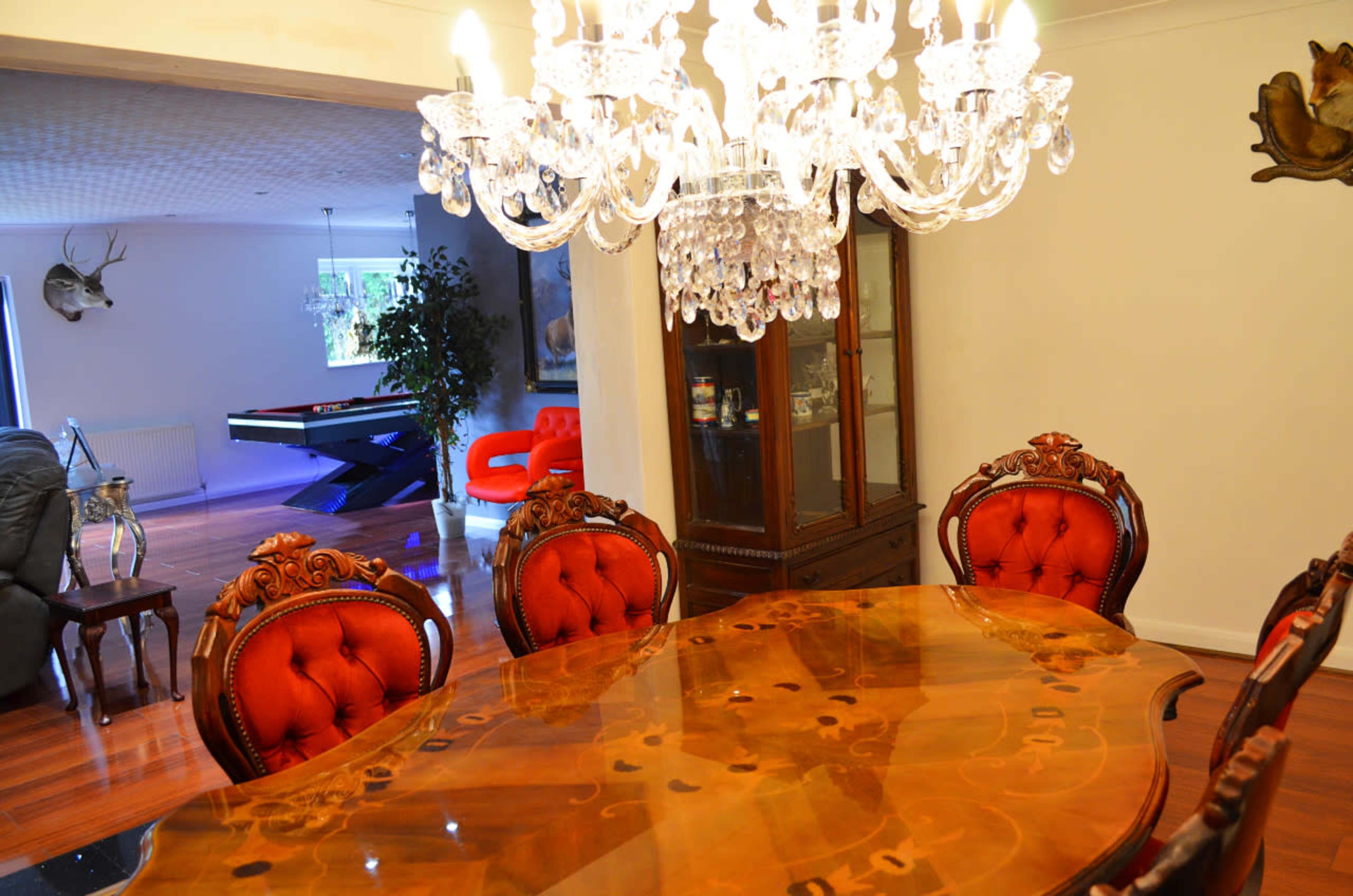 A crystal chandelier hangs above an ornate wooden dining table surrounded by upholstered chairs, with a view of a modern living space in the background.