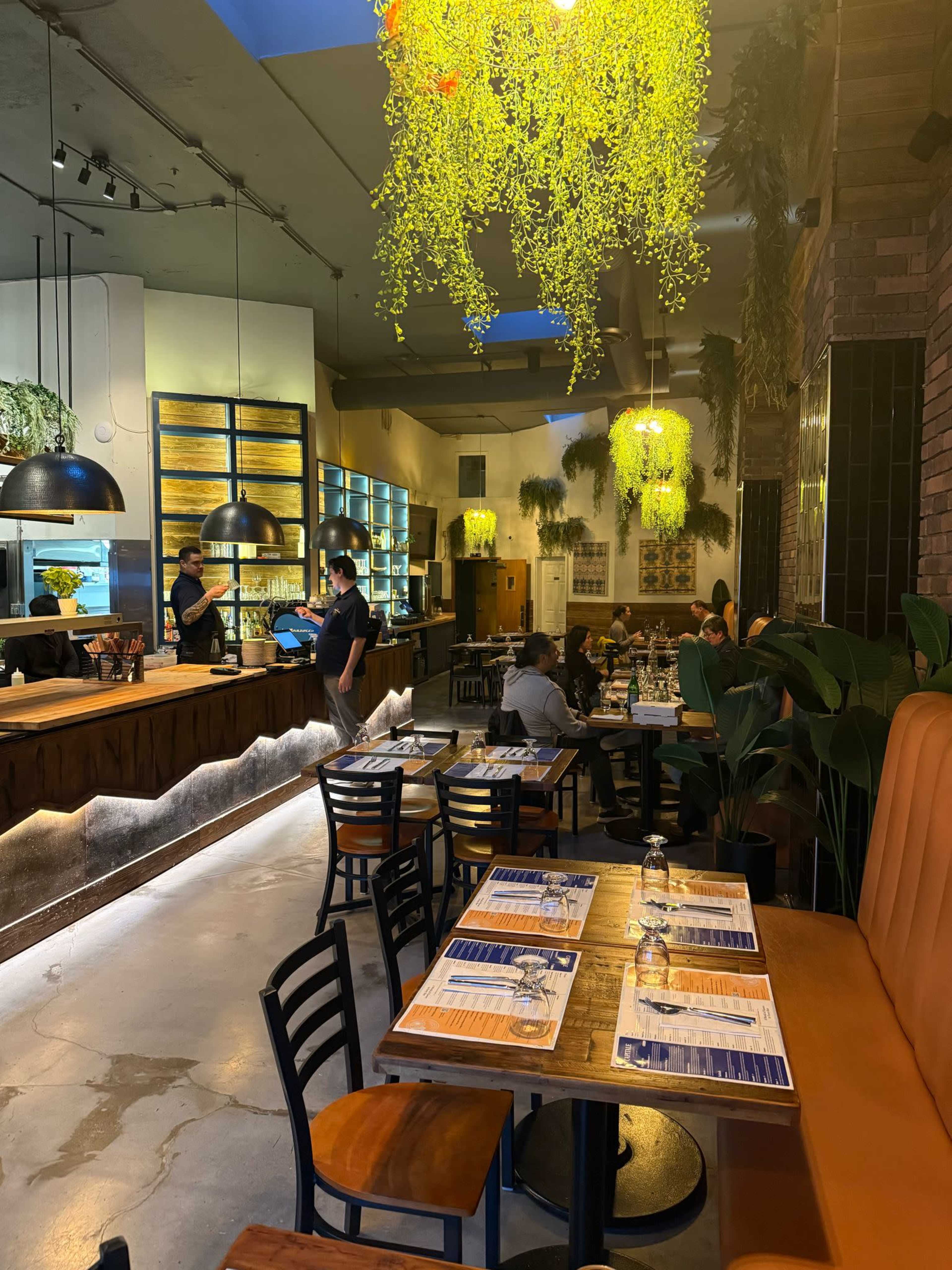 The image shows a modern restaurant interior with wooden tables, black chairs, and hanging greenery, featuring patrons dining and staff attending to customers at the bar area.