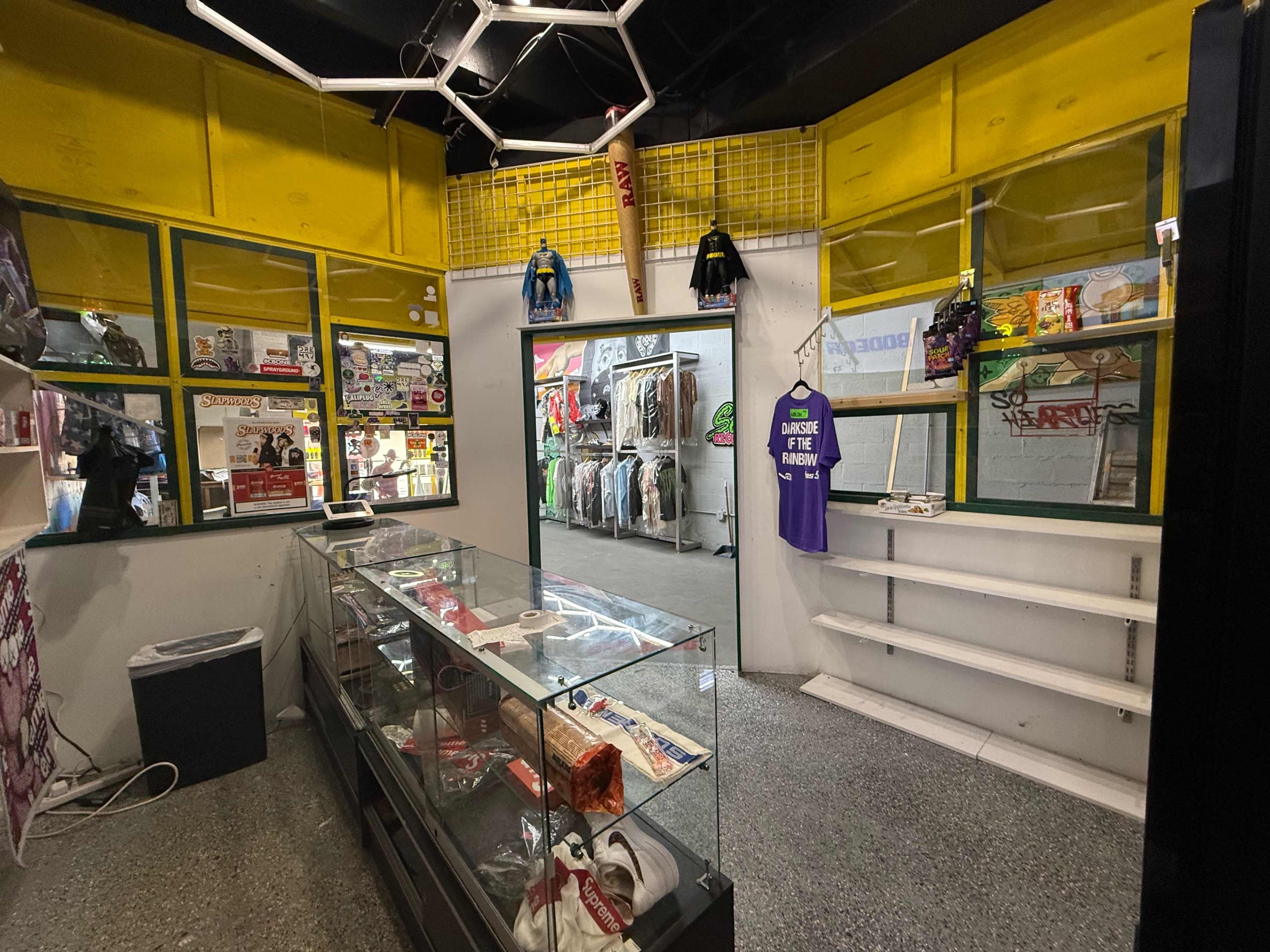 The image shows the interior of a retail store featuring a glass display case, shelves stocked with various items, and clothing hung on racks along the walls.