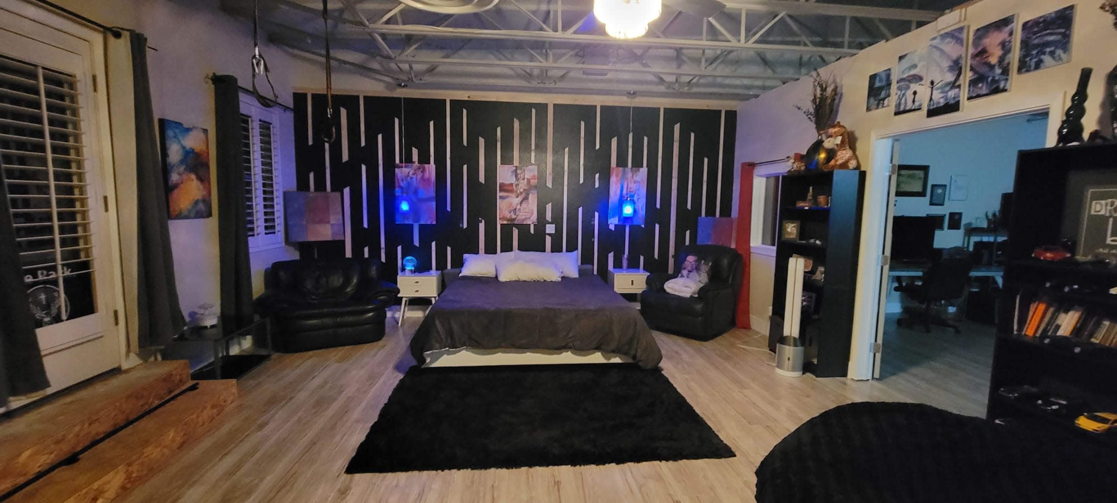 The image shows a spacious room featuring a large bed in the center, dark furniture, and blue ambient lighting, with a workspace visible in the background.