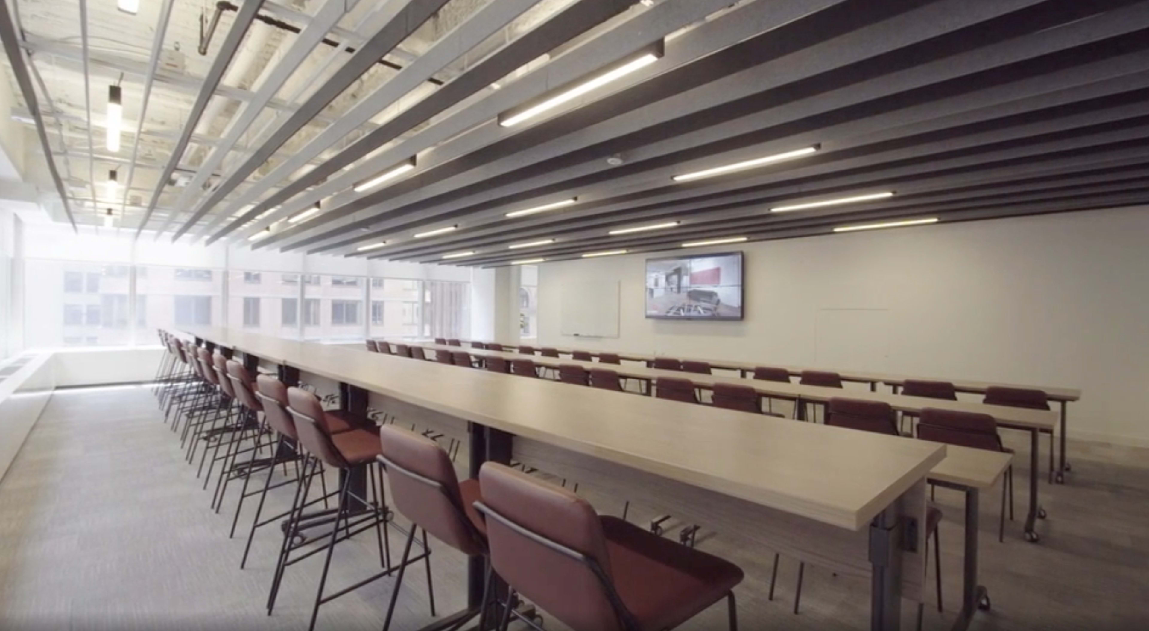 The image shows a modern meeting room with a long table, rows of chairs, and a large screen on the wall.