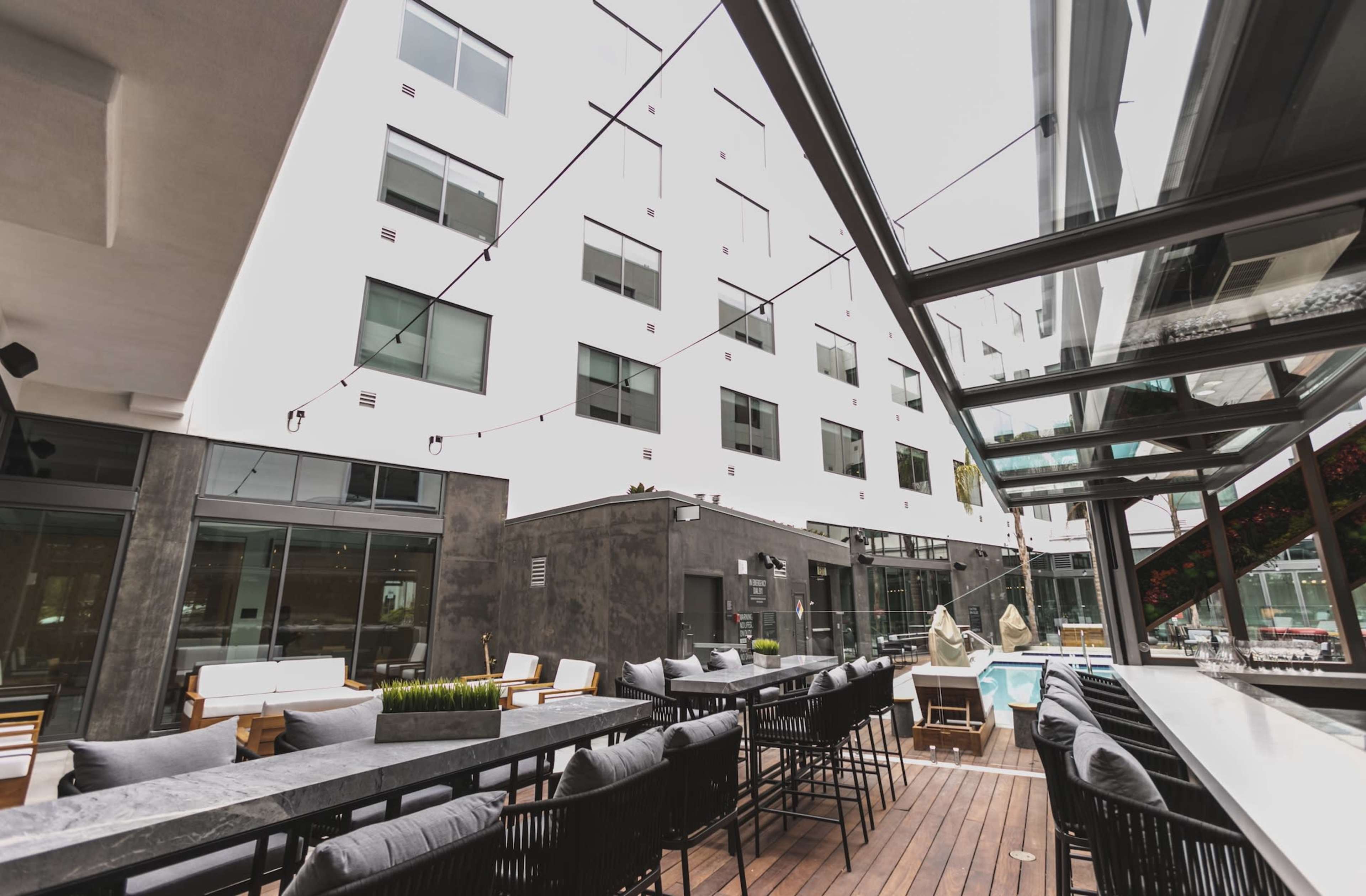 The image shows a modern outdoor dining area with black bar stools and a long table, surrounded by a contemporary building with large windows.