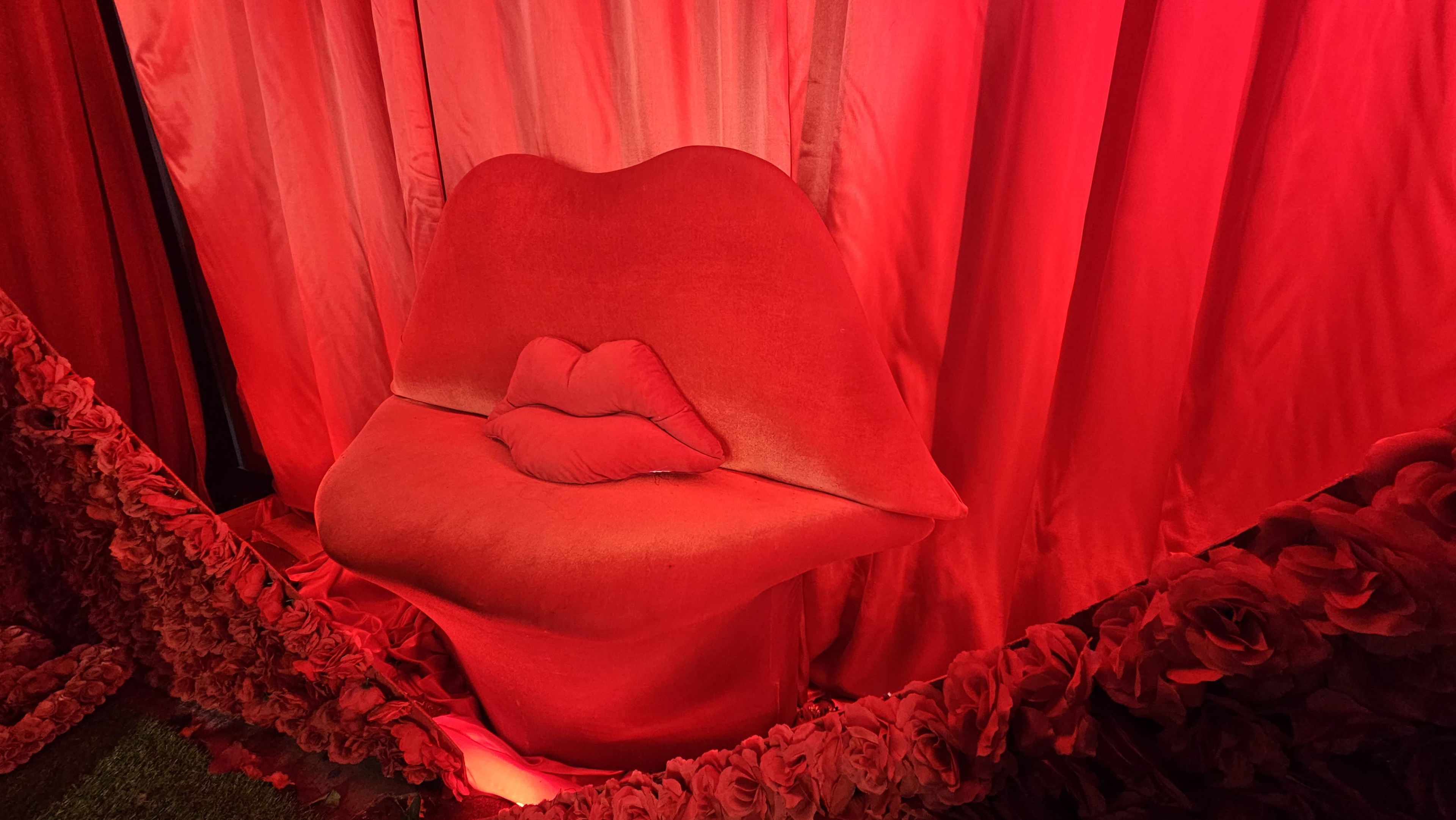 A red, lip-shaped chair sits against a backdrop of matching red curtains and floral decorations.