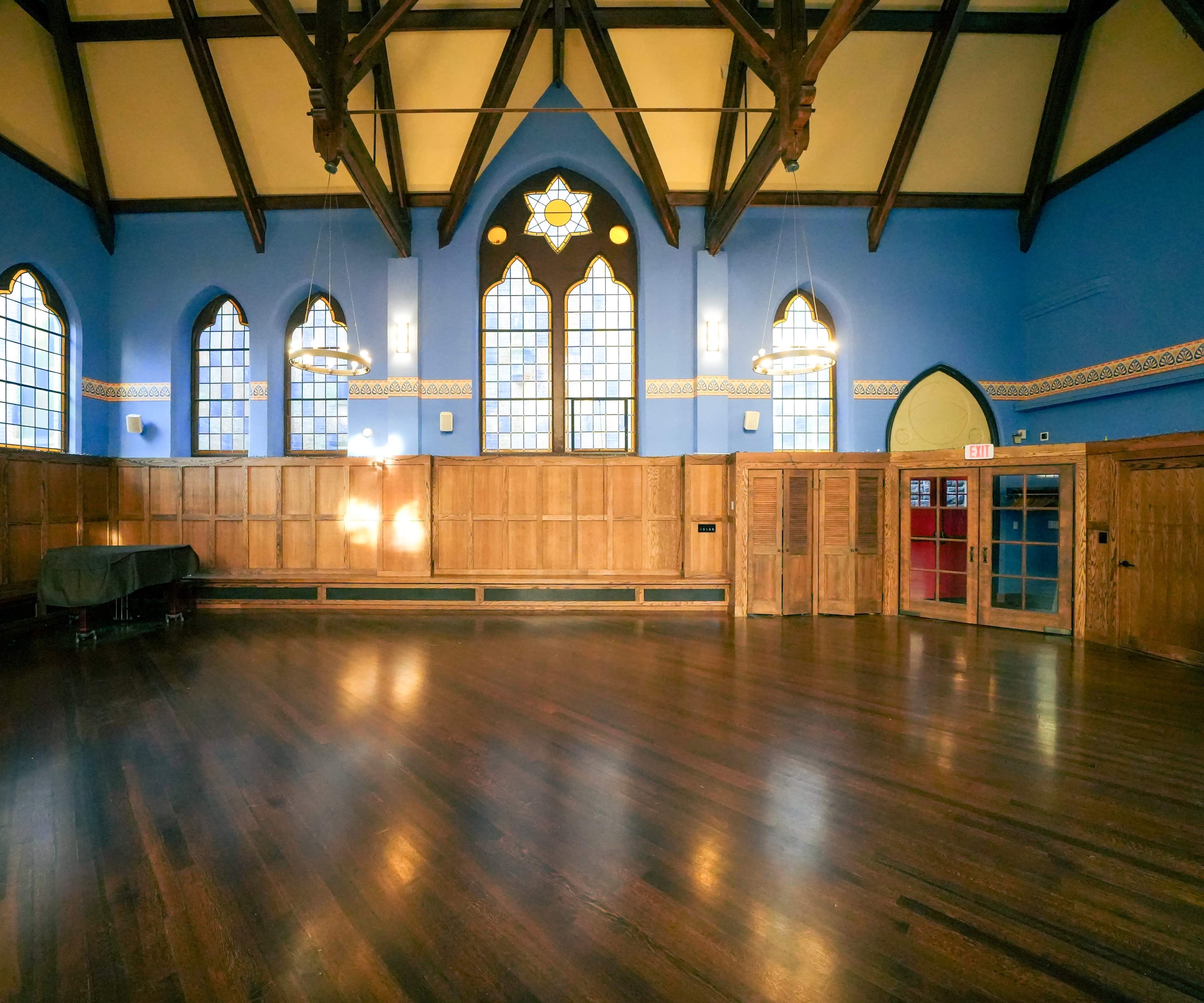 A spacious interior room with wooden floors, high ceilings, and large windows featuring stained glass, illuminated by overhead lights.