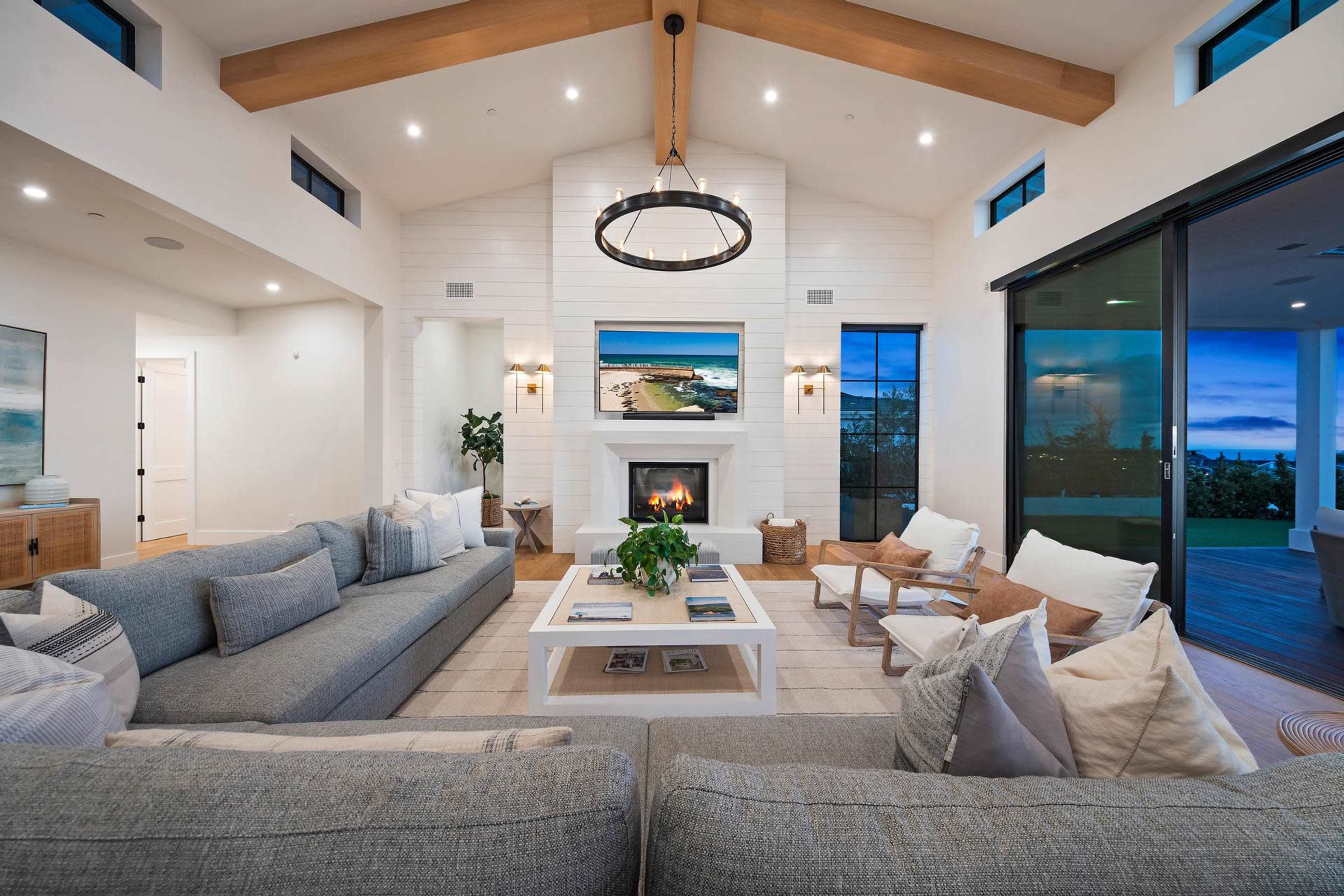 The image shows a modern living room featuring a large sectional sofa, a coffee table in the center, a fireplace with a mounted television above it, and glass doors leading to an outdoor area.