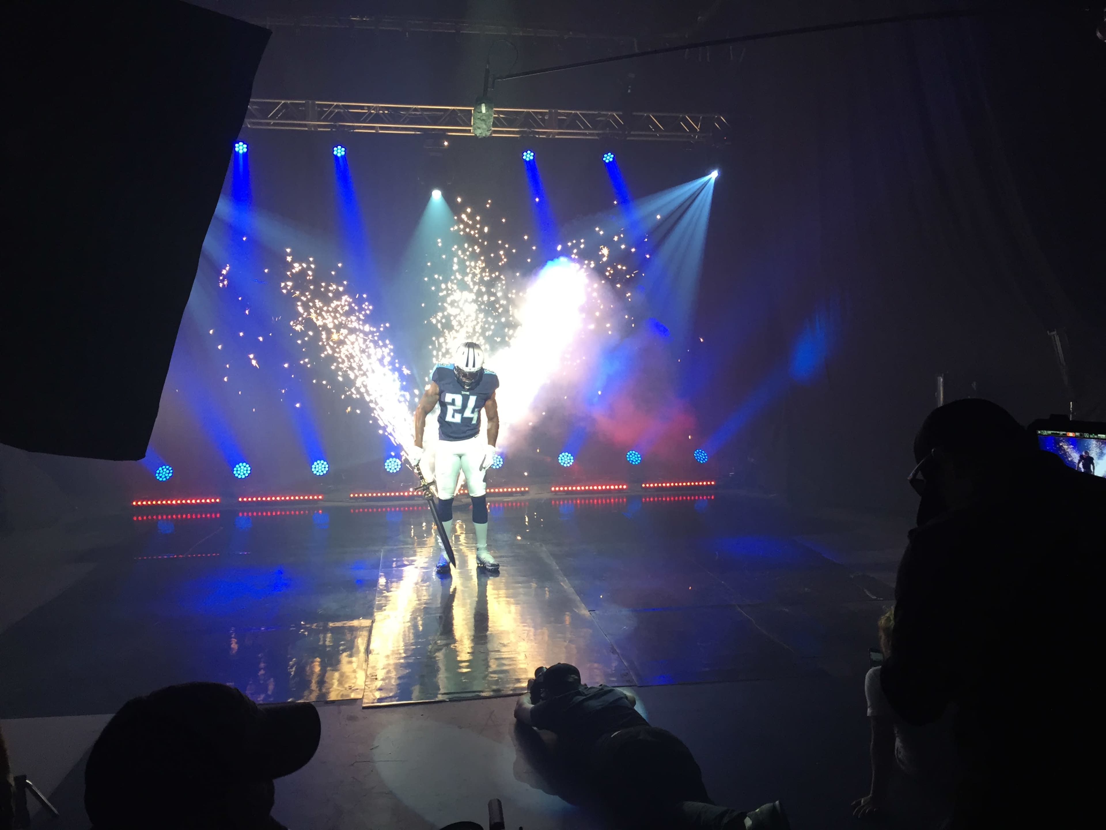 A football player in a blue jersey with the number 24 stands on a reflective stage illuminated by bright lights and sparks.