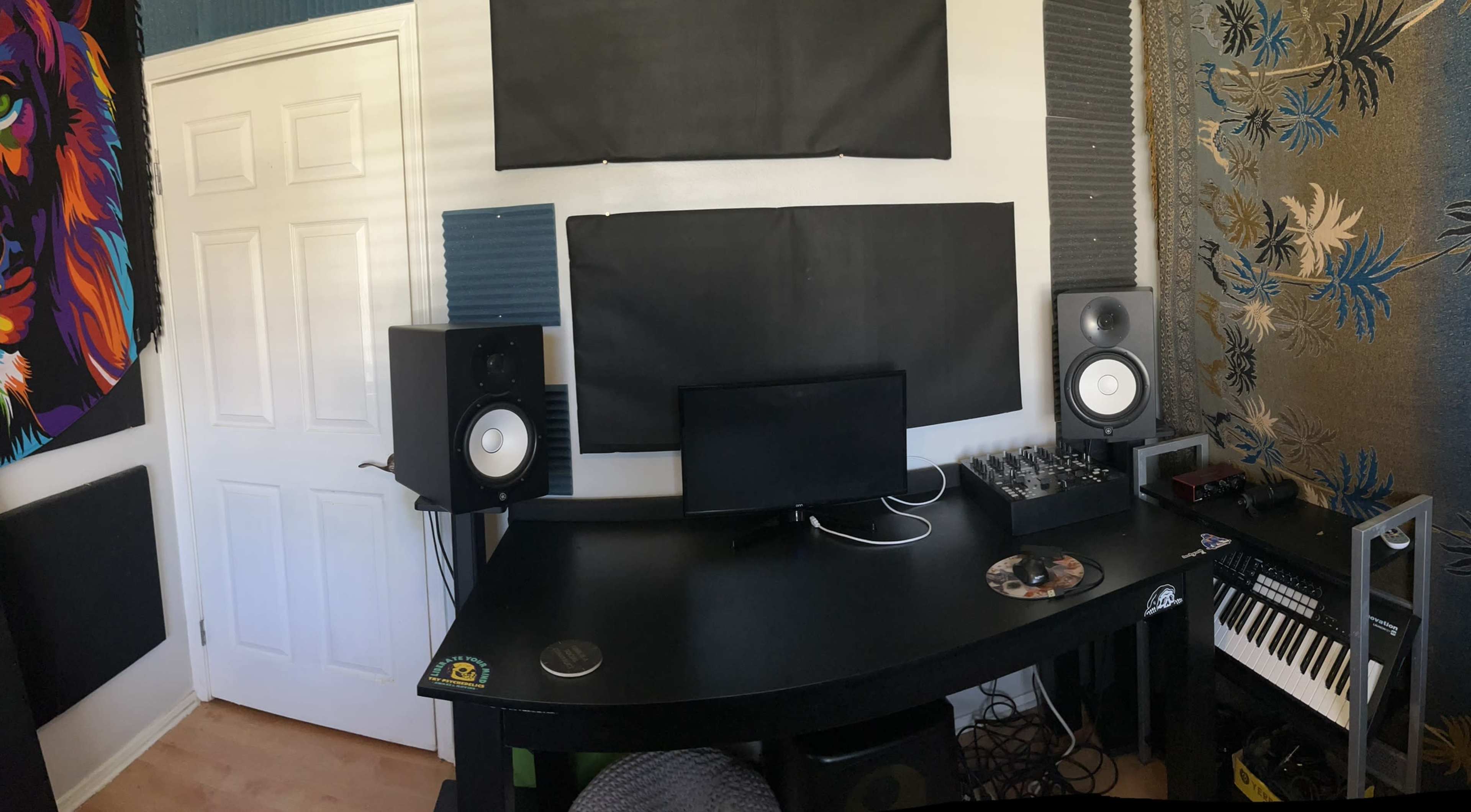 The image shows a music production workspace featuring a black desk with a computer monitor, audio interface, speakers, and a keyboard, surrounded by acoustic panels on the walls.