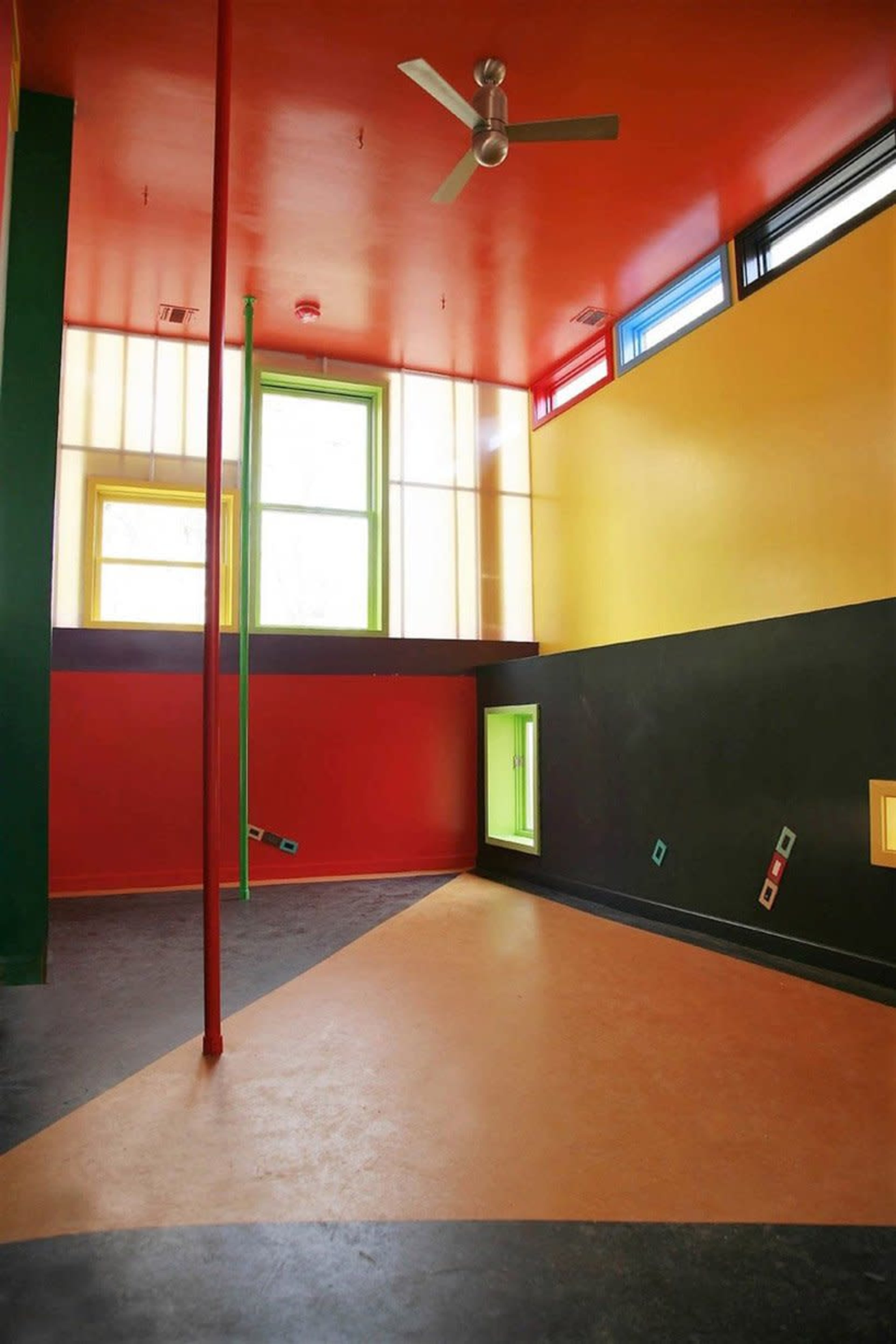 The image shows a brightly colored interior room featuring red, yellow, green, and black walls, with windows and a ceiling fan.