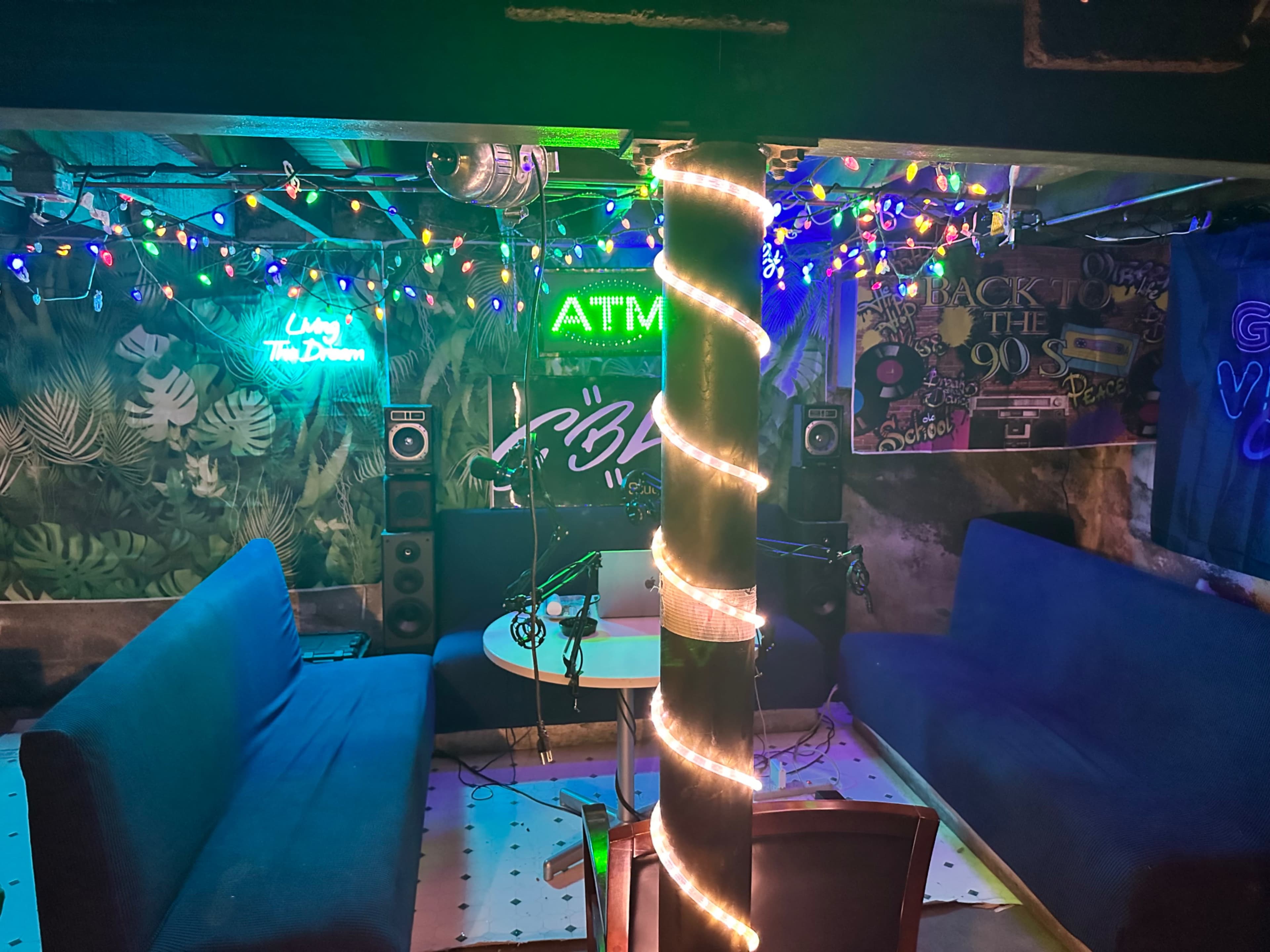 A dimly lit lounge area with colorful string lights, featuring two sofas, a table, and speakers against a decorated wall.