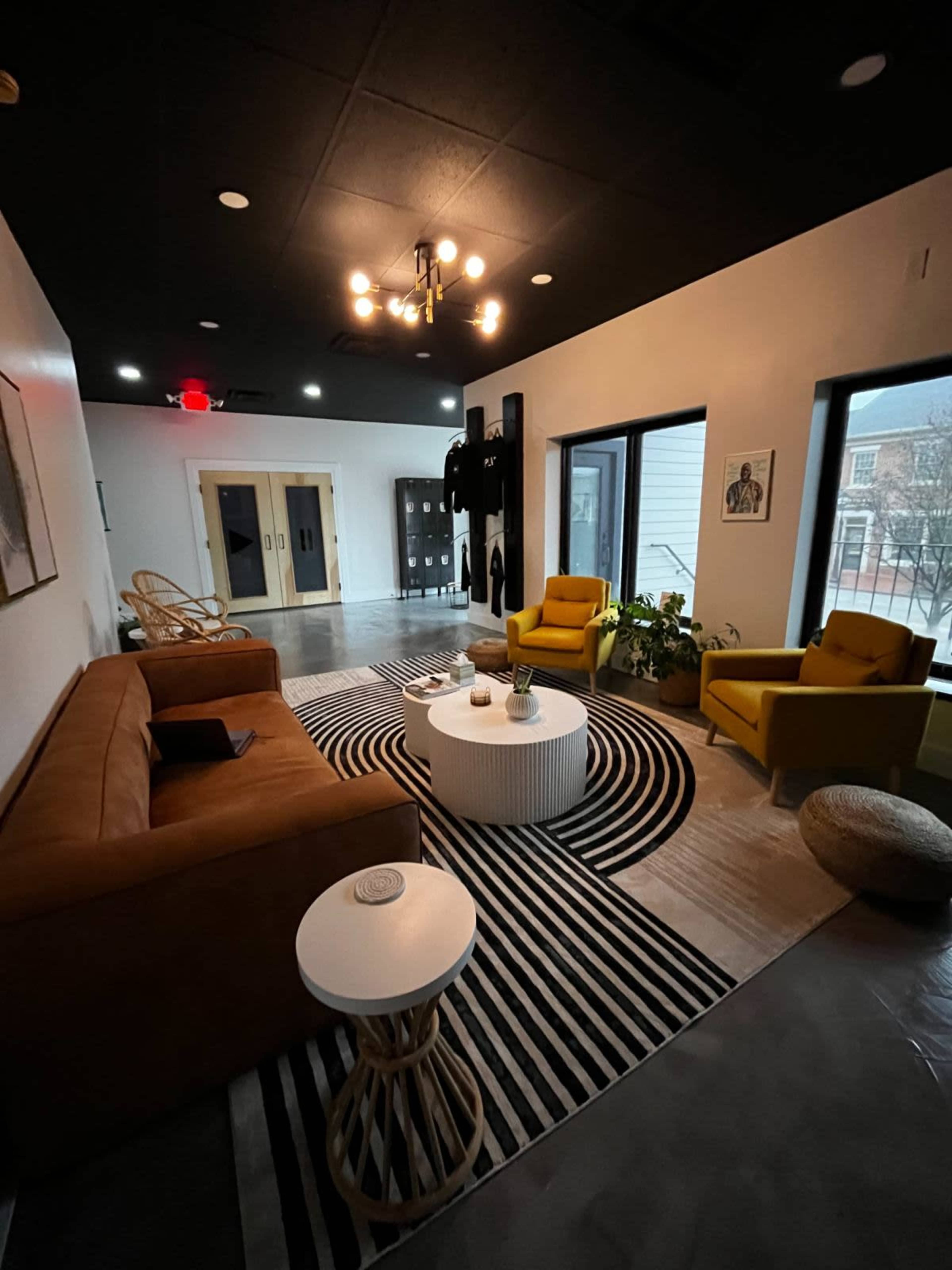 A modern lounge area with a brown sofa, yellow armchairs, a round coffee table, and a patterned rug, featuring plants and artwork on the walls.
