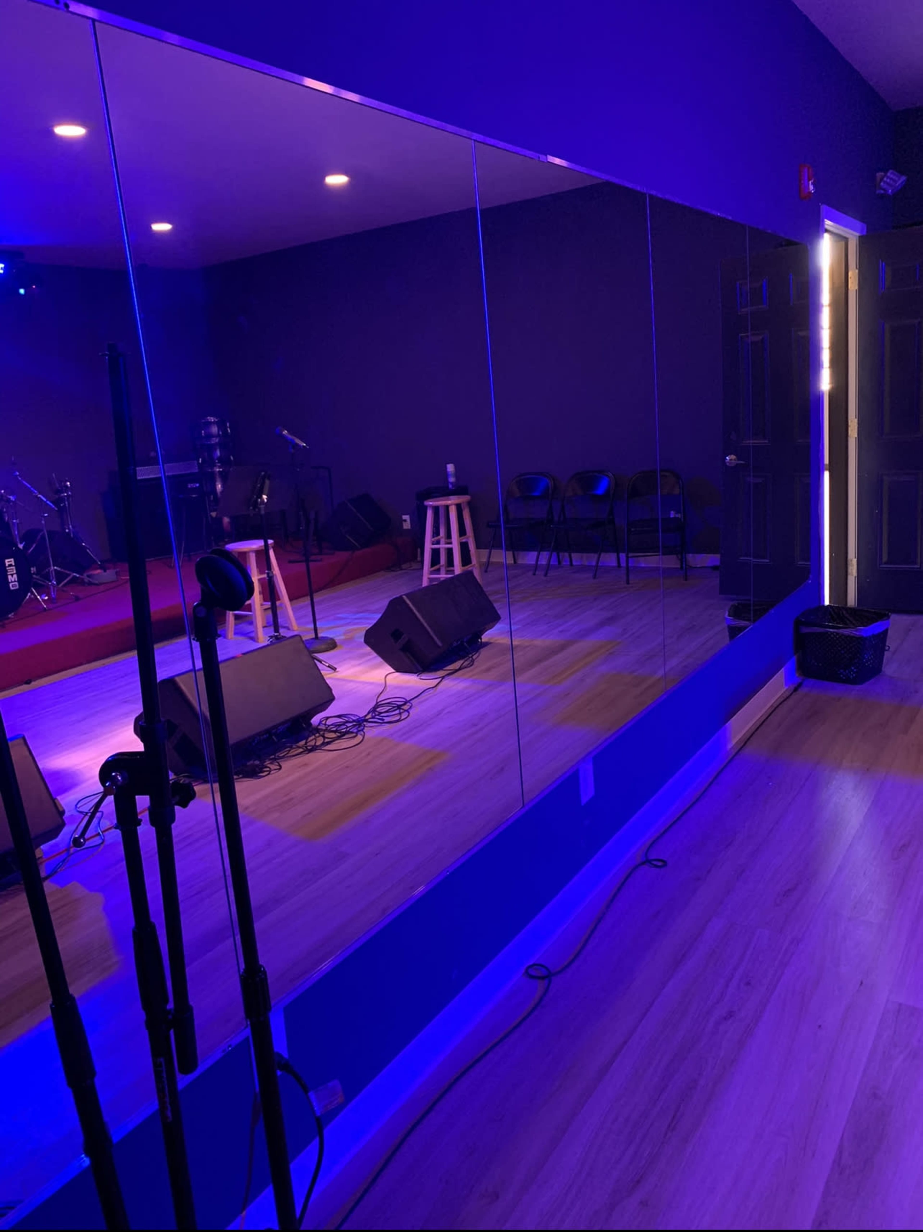 The image shows a small stage with musical equipment, surrounded by seating, under blue lighting, and reflected in a large mirror.