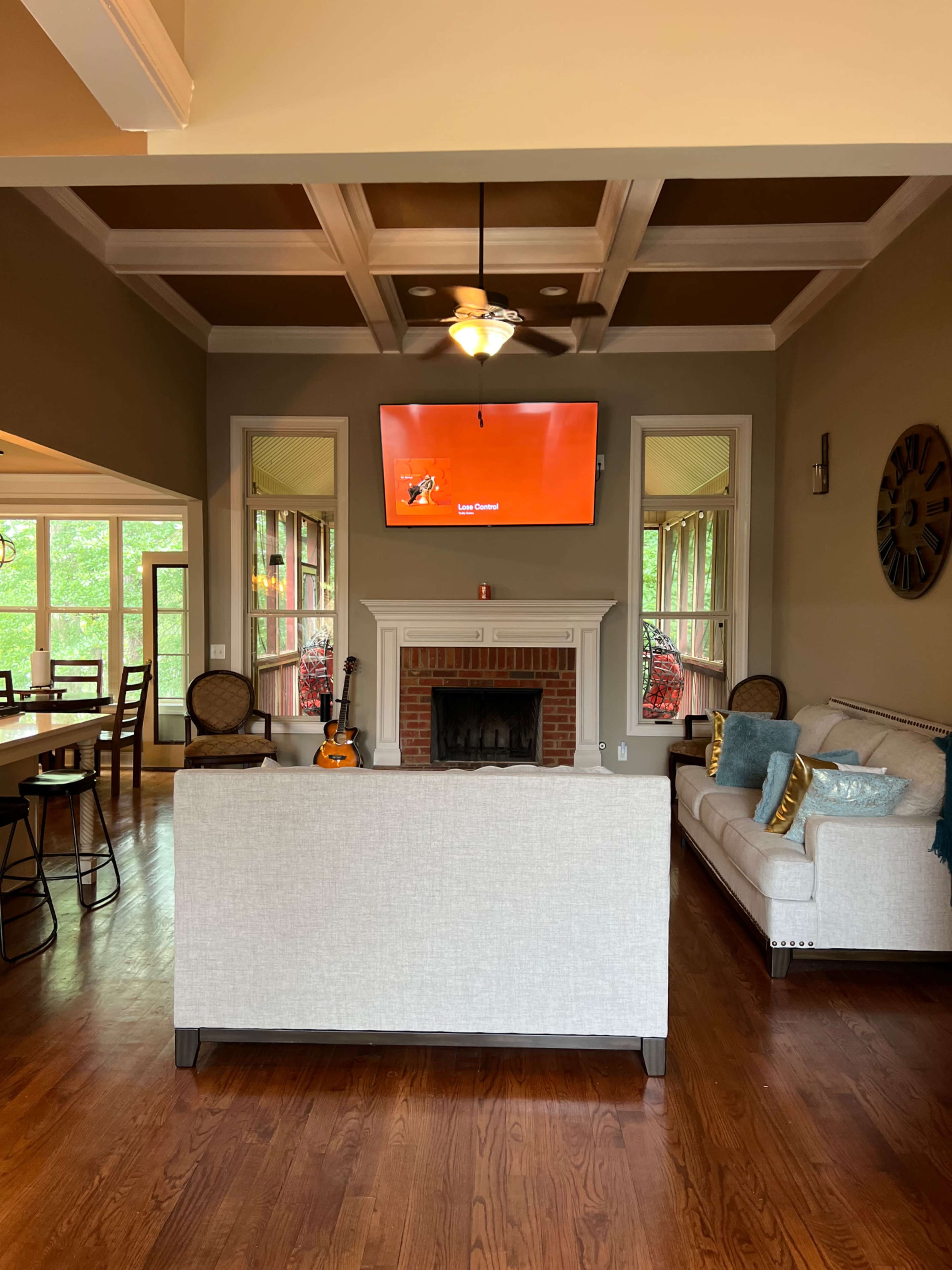 The image shows a cozy living room featuring a white sofa, a fireplace with a television mounted above it, and large windows that open to a screened porch.