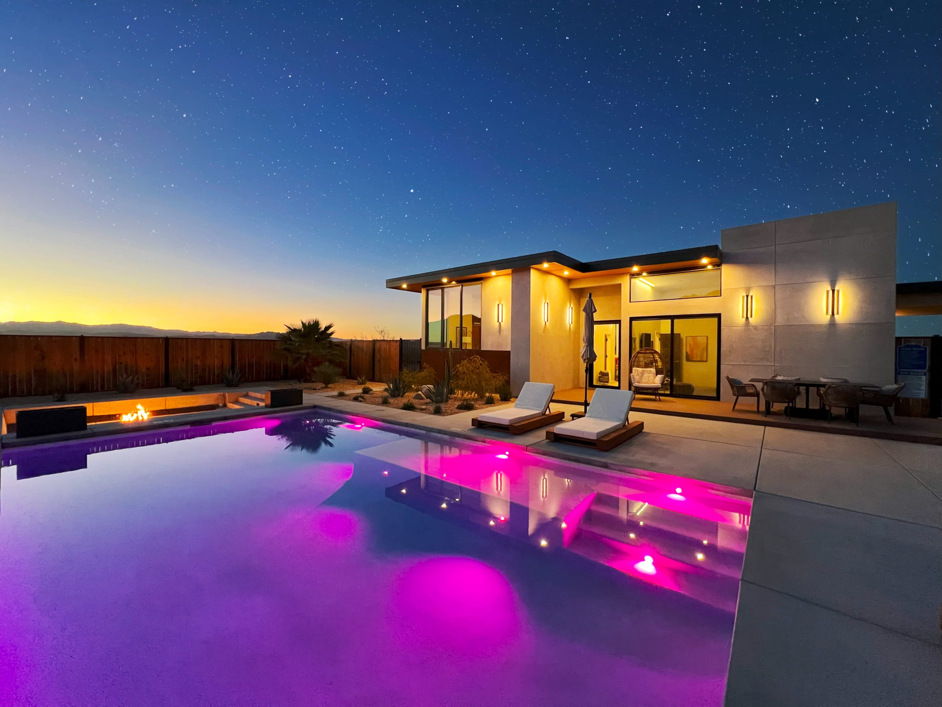 A modern home with a pool illuminated in purple sits under a twilight sky filled with stars.