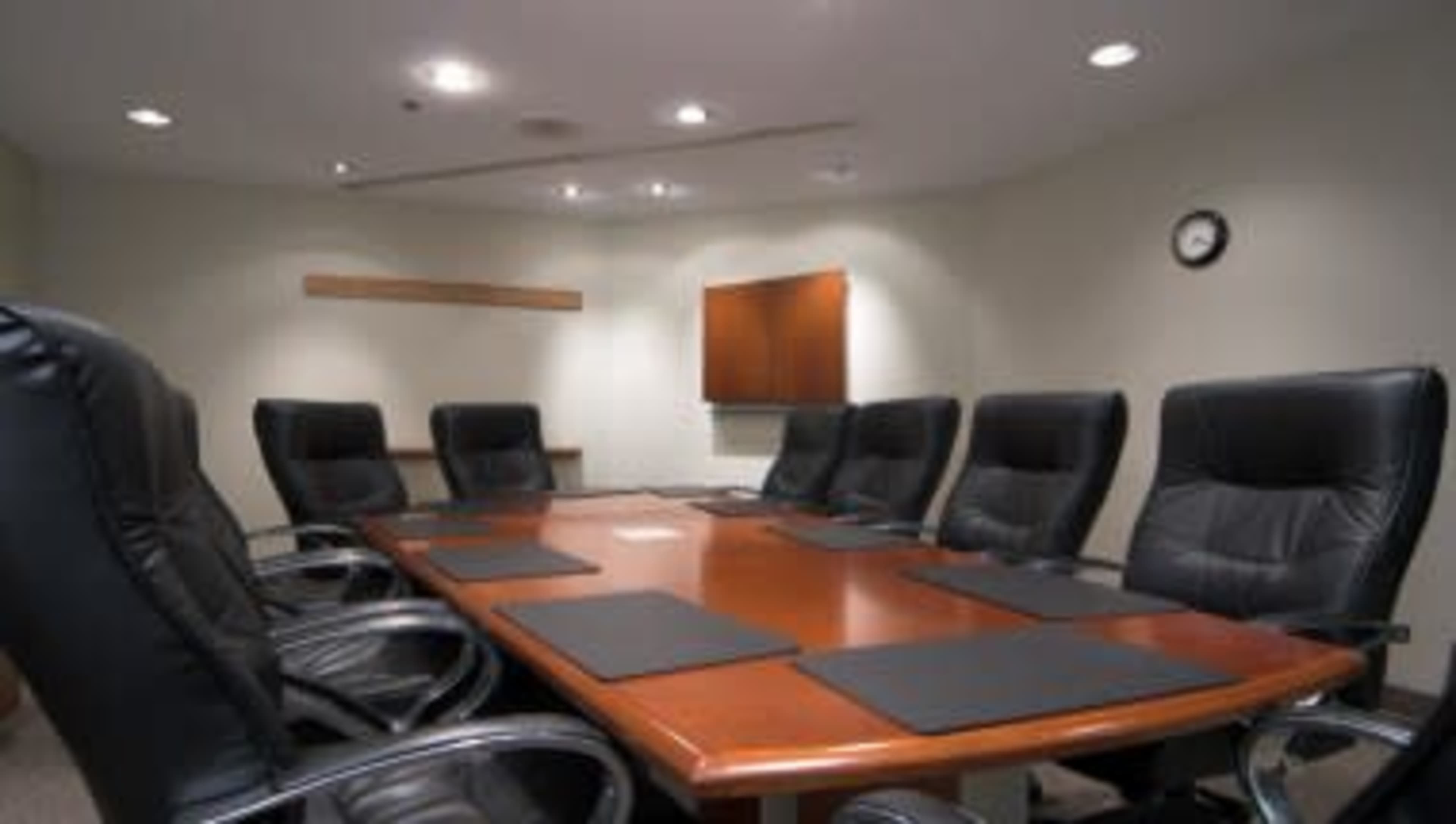 A large conference room features a wooden table surrounded by eight black leather chairs, with a clock and a television mounted on the wall.