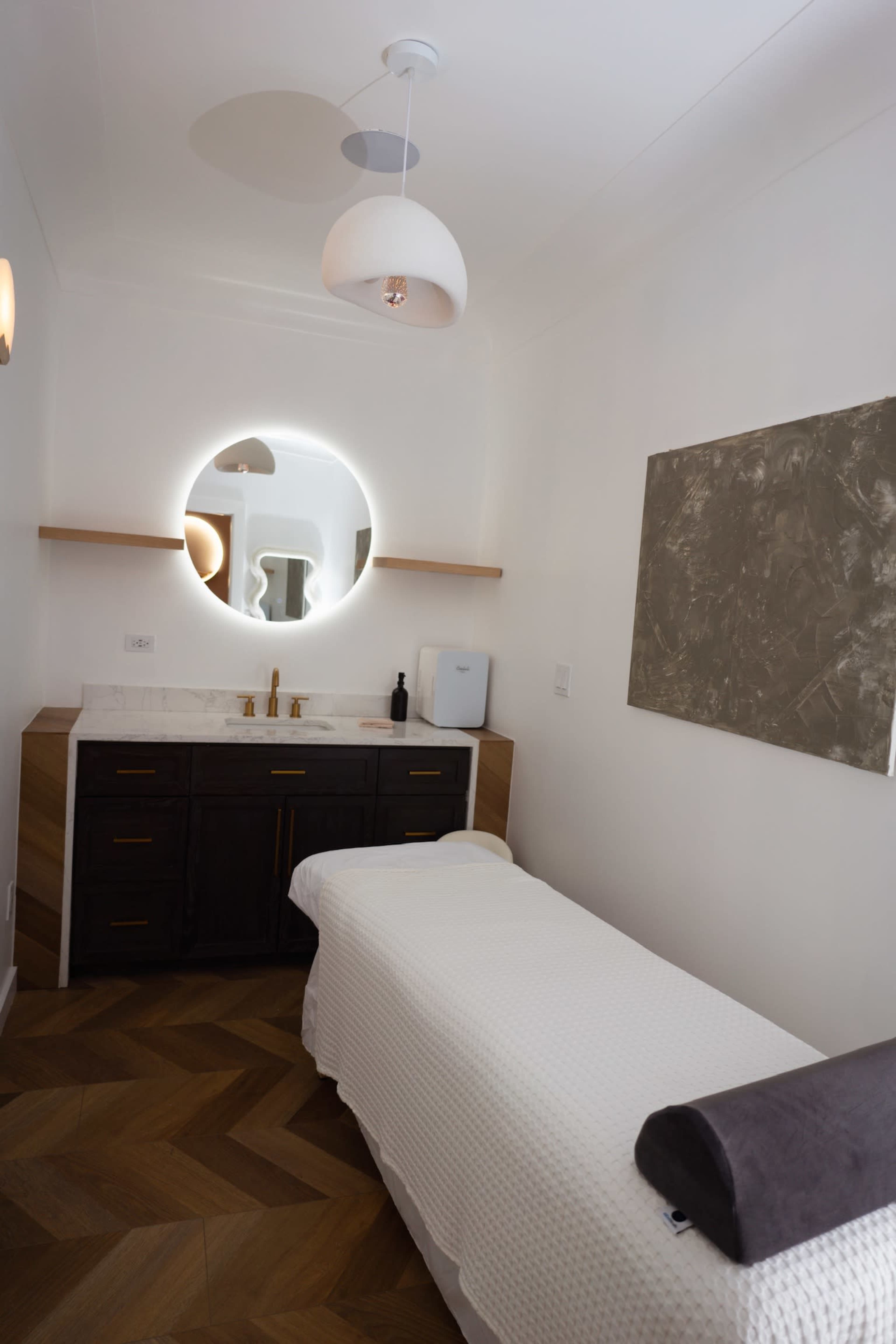 A minimalist treatment room with a massage table, a round mirror, and a dark wood cabinet against a light-colored wall.