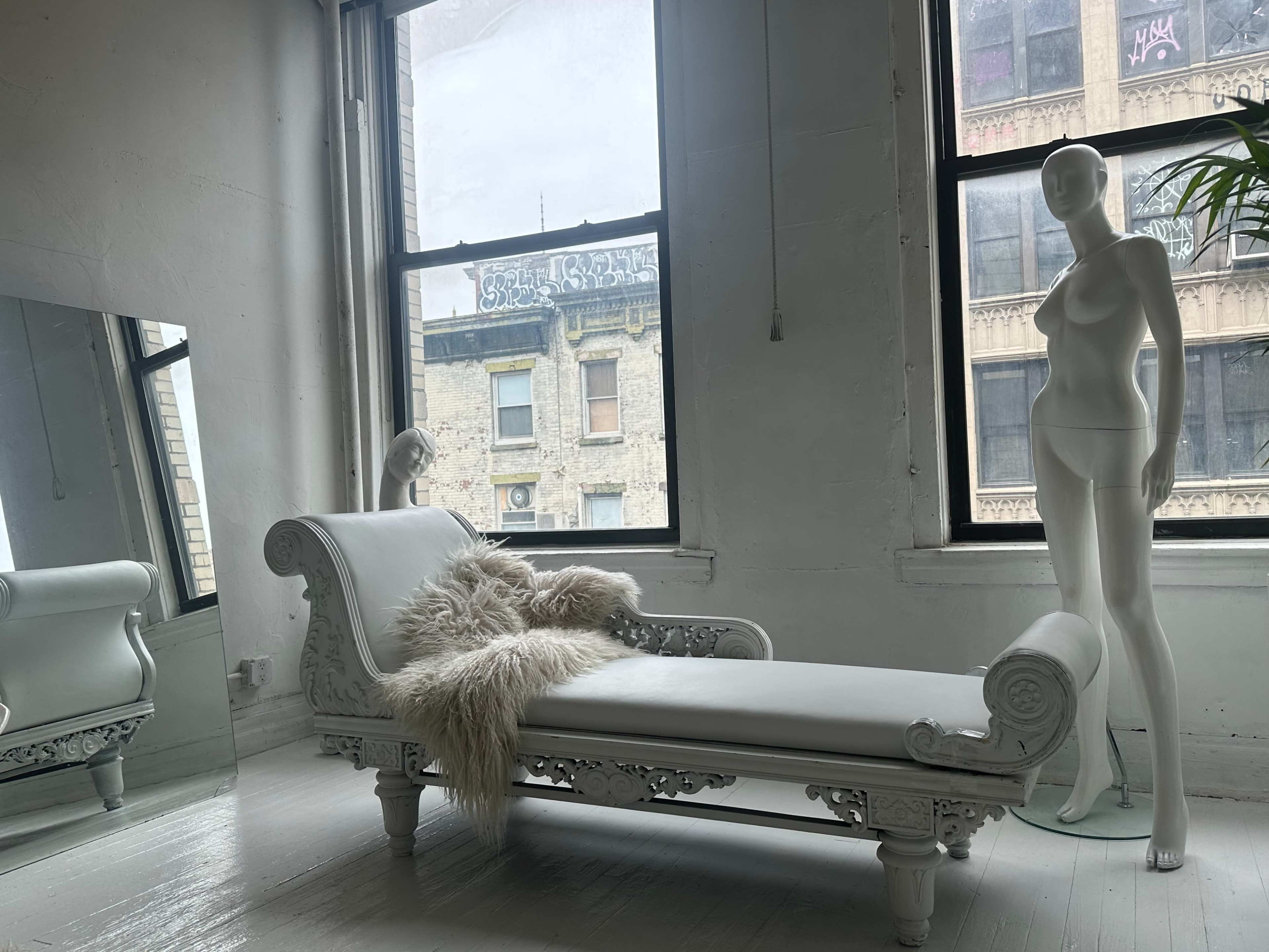 The image shows a white chaise lounge with a fur throw, a full-length mirror, and a mannequin standing next to a large window overlooking a city street.