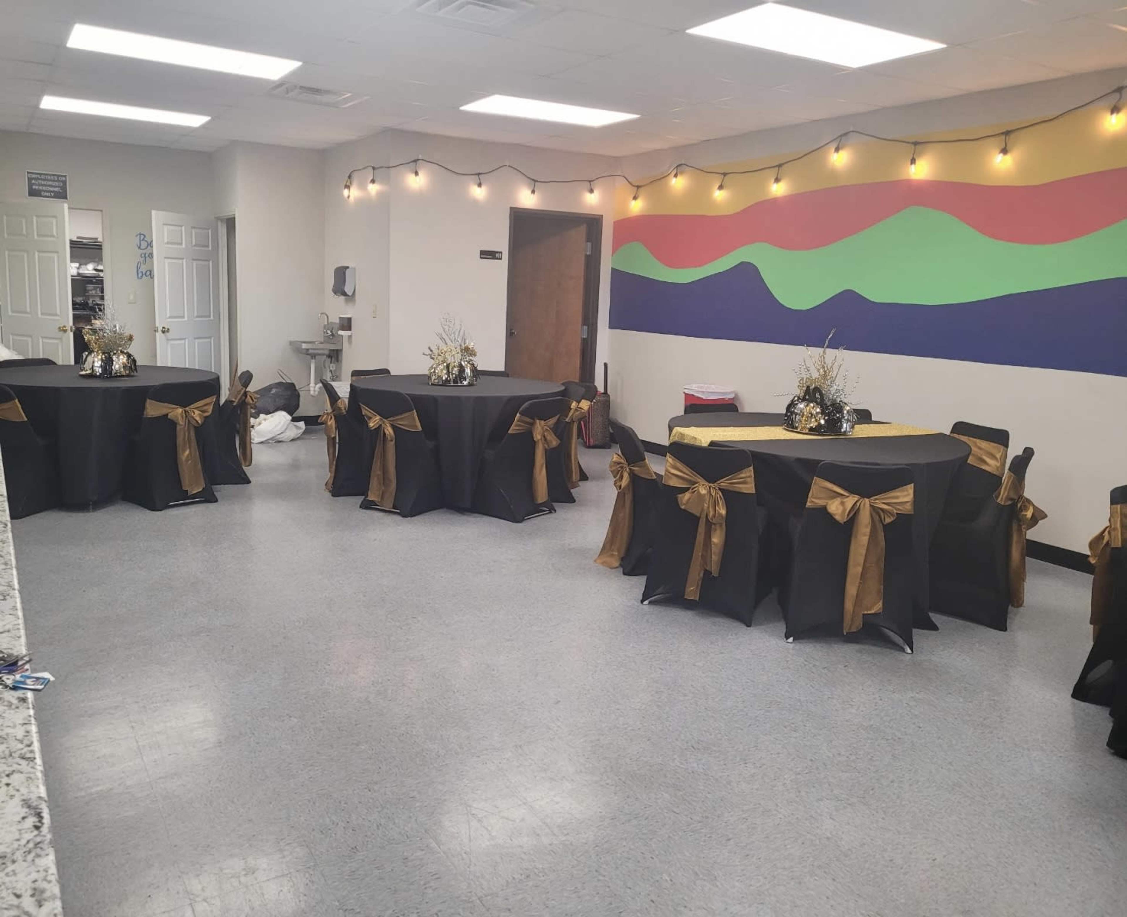 The image shows a room set up for an event, featuring several black tables adorned with gold bows and decorative centerpieces, illuminated by string lights on the walls.