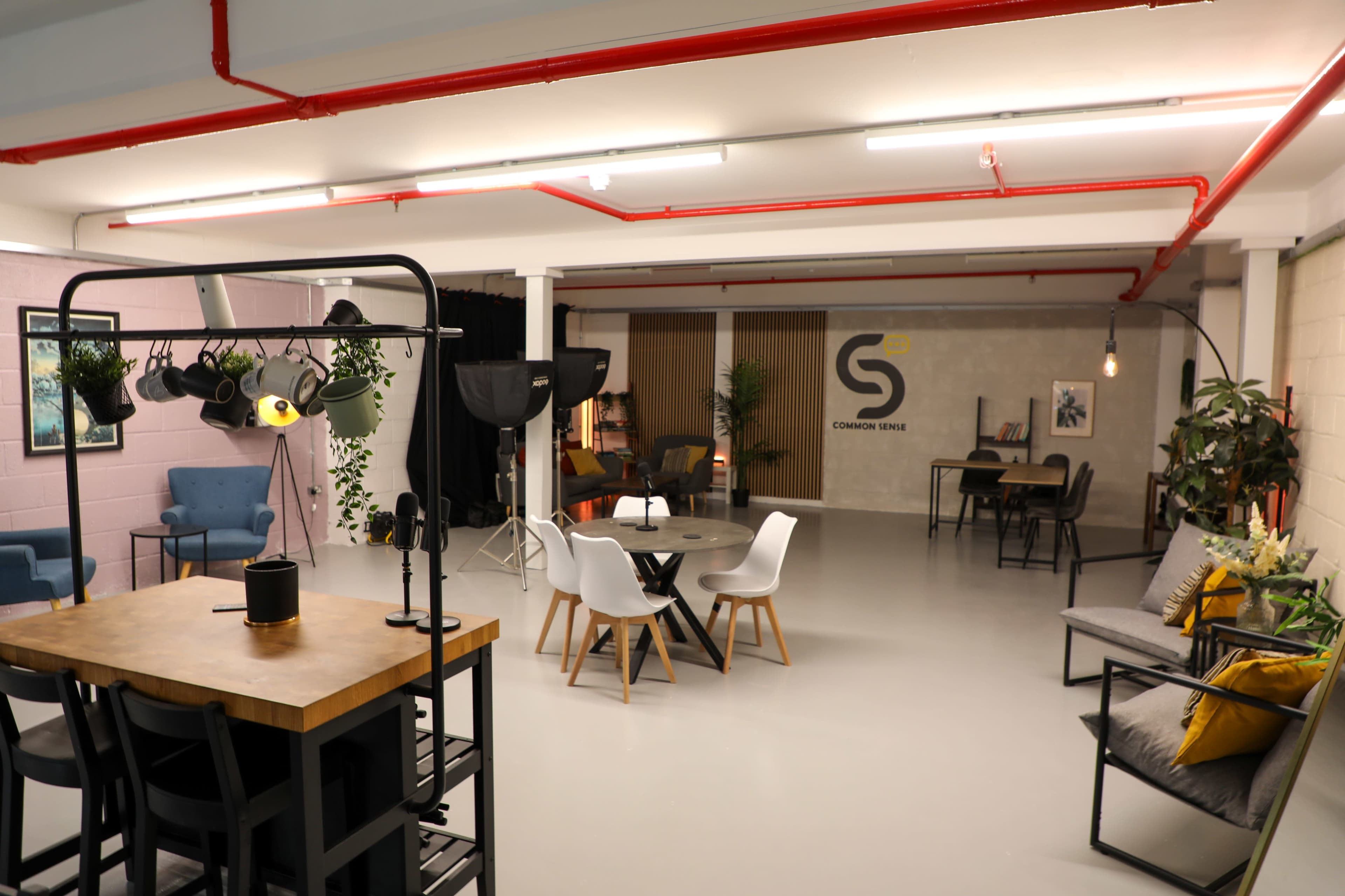 The image shows a bright, modern co-working space featuring various seating arrangements, tables, and decorative plants.