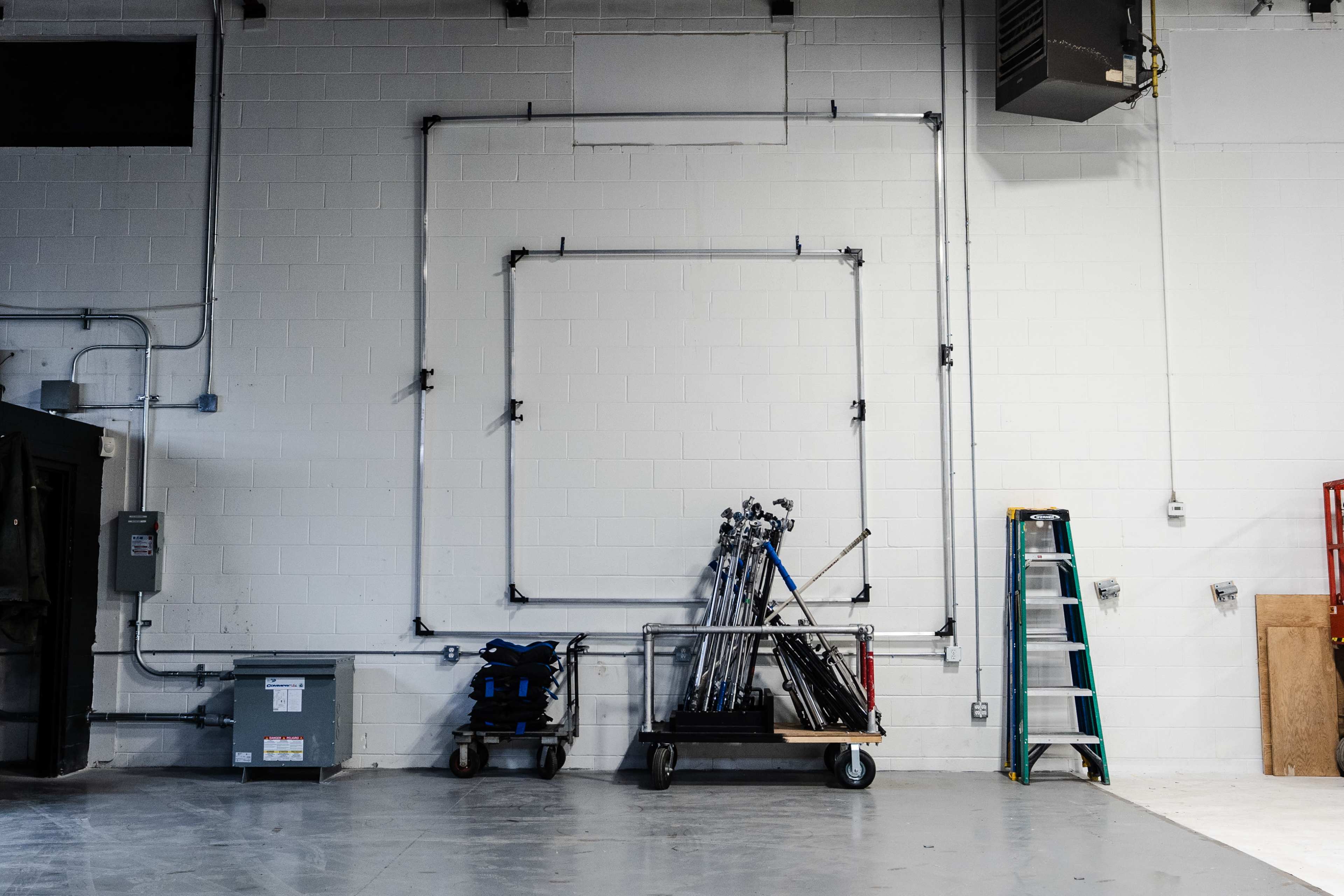 A wall in an industrial space with pipes forming a rectangular outline and various equipment, including tripods and a ladder, arranged on a cart.
