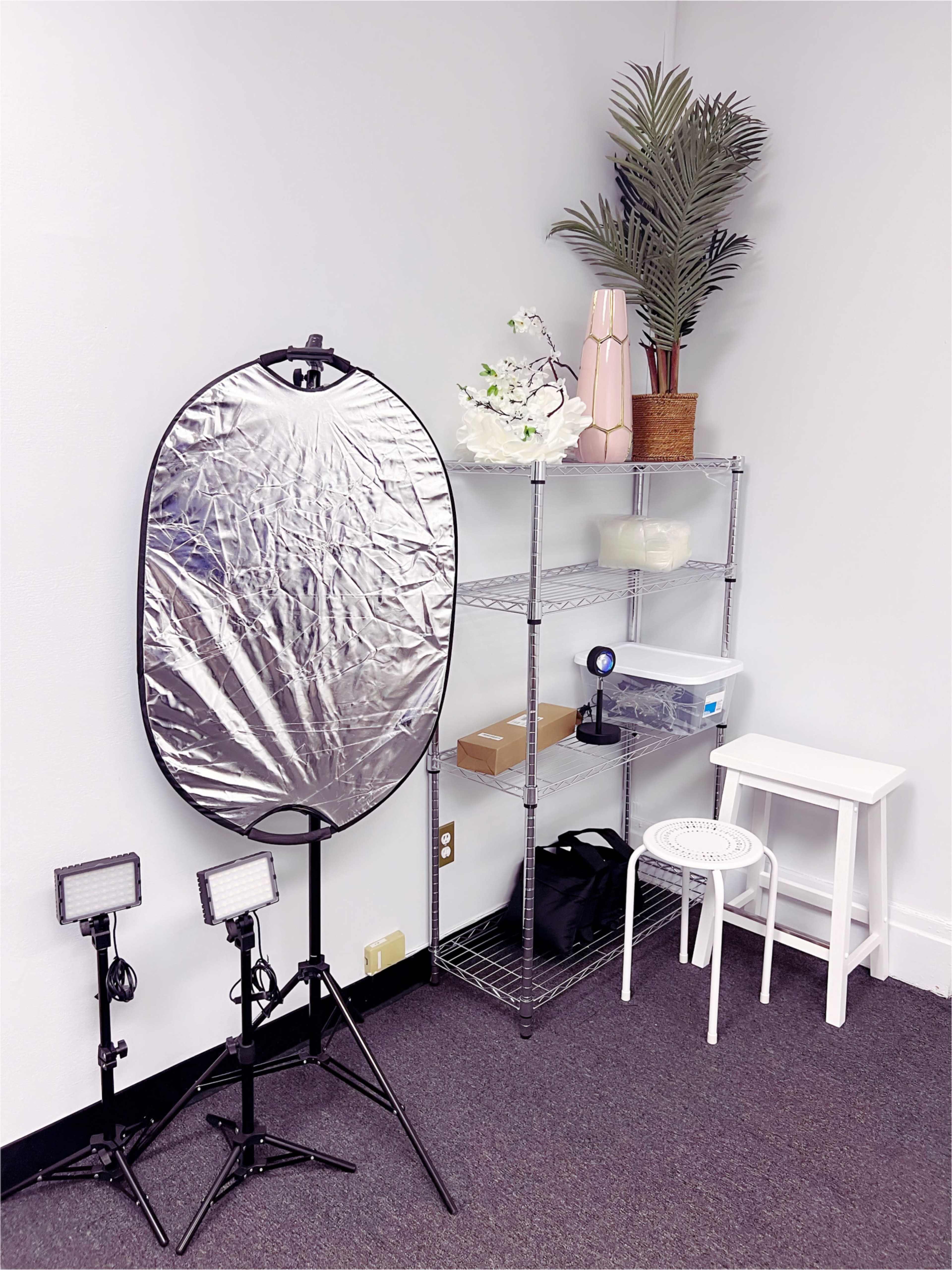 The image shows a corner setup with a silver reflector on a stand, two lighting fixtures, a metal shelving unit with various items, and a small white stool.