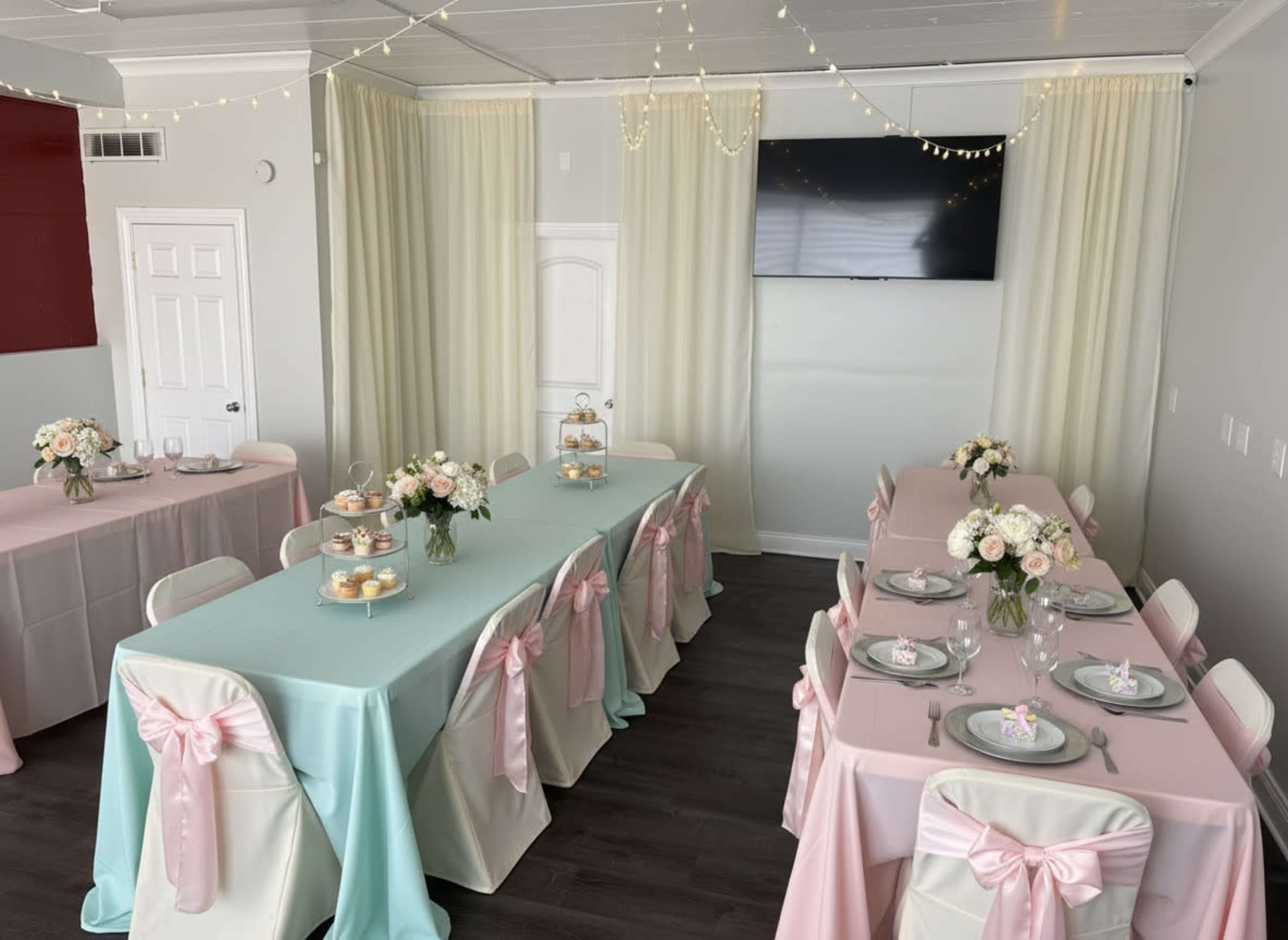 The image shows a decorated event space with two tables covered in pastel tablecloths, one in mint green and the other in light pink, adorned with floral centerpieces and matching chair sashes.