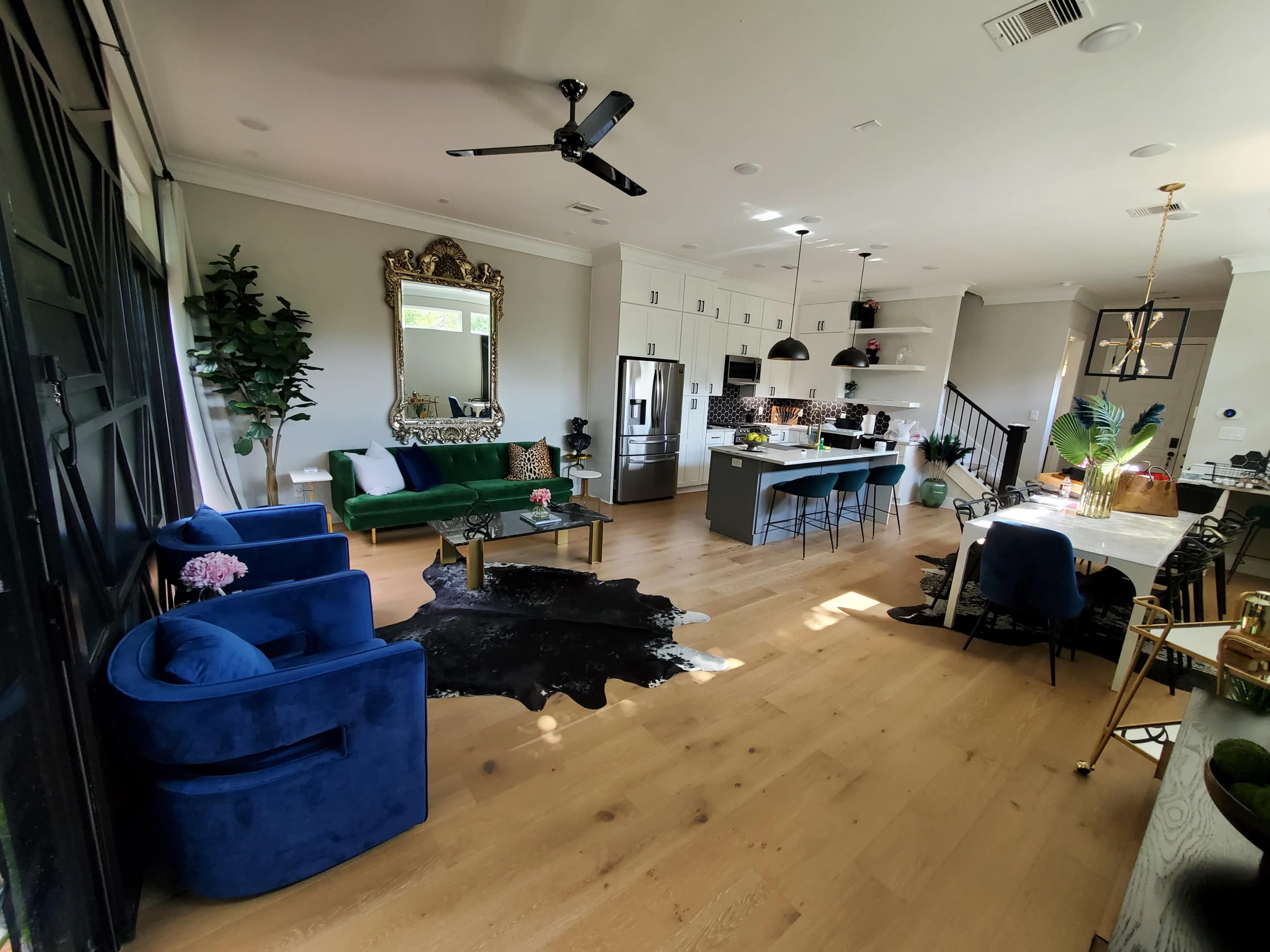 A spacious living area features a green velvet sofa, blue chairs, a large dining table, and an open kitchen with modern appliances.