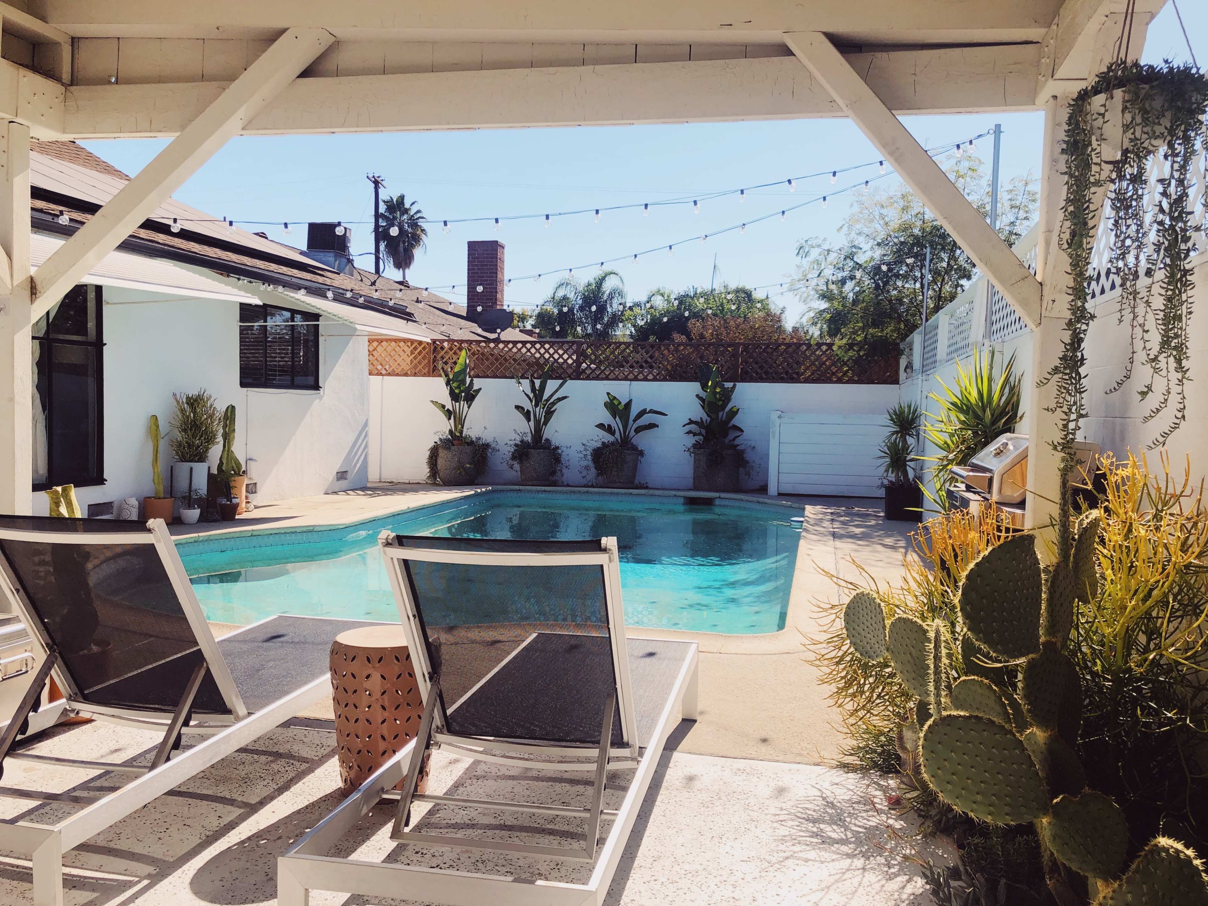 A backyard pool area features two lounge chairs by the water, surrounded by potted plants and a wall-mounted information board.