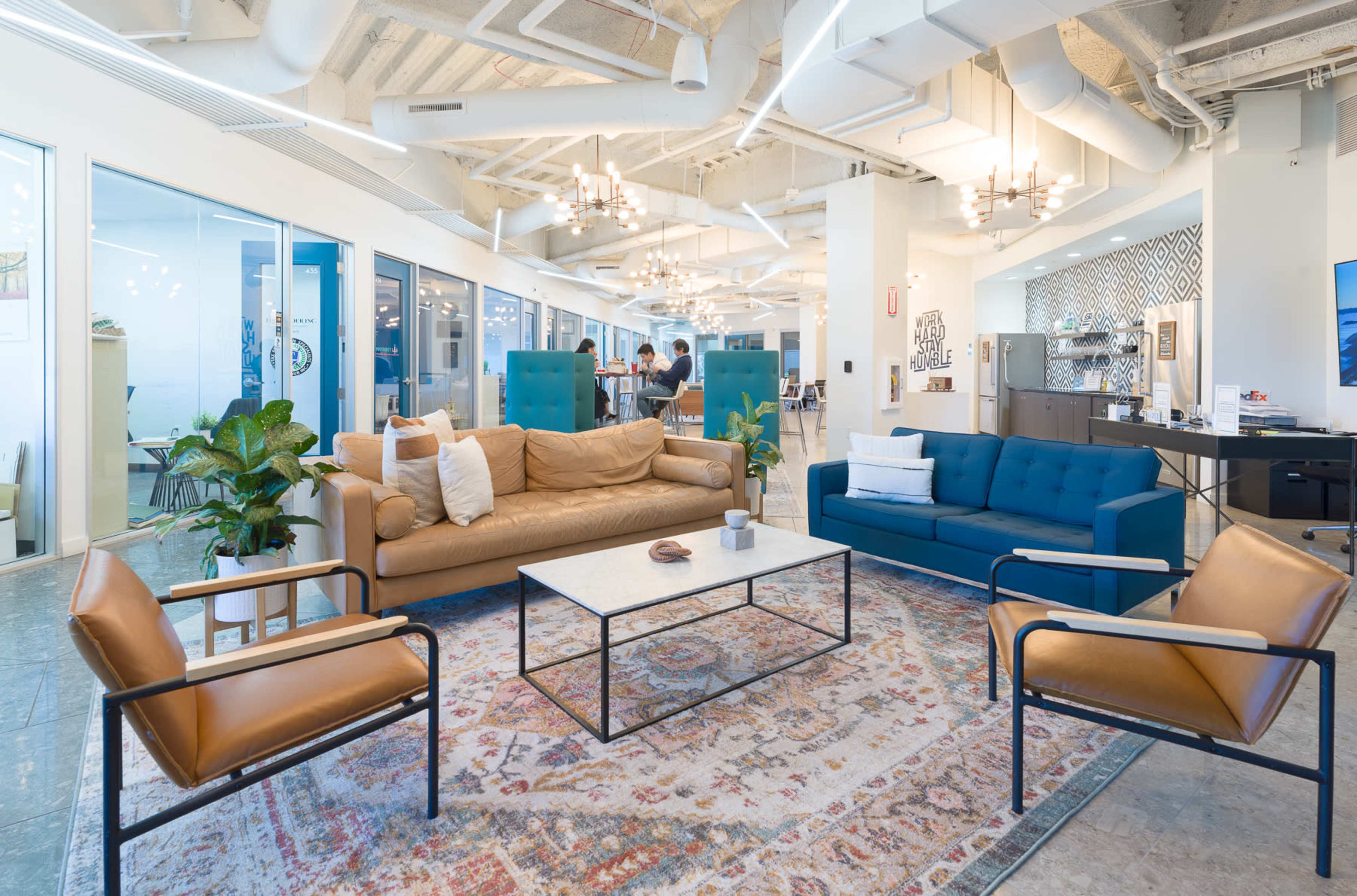 A modern office space features a mix of seating, including a brown leather sofa, a blue tufted couch, and several armchairs, arranged around a coffee table on a decorative rug.