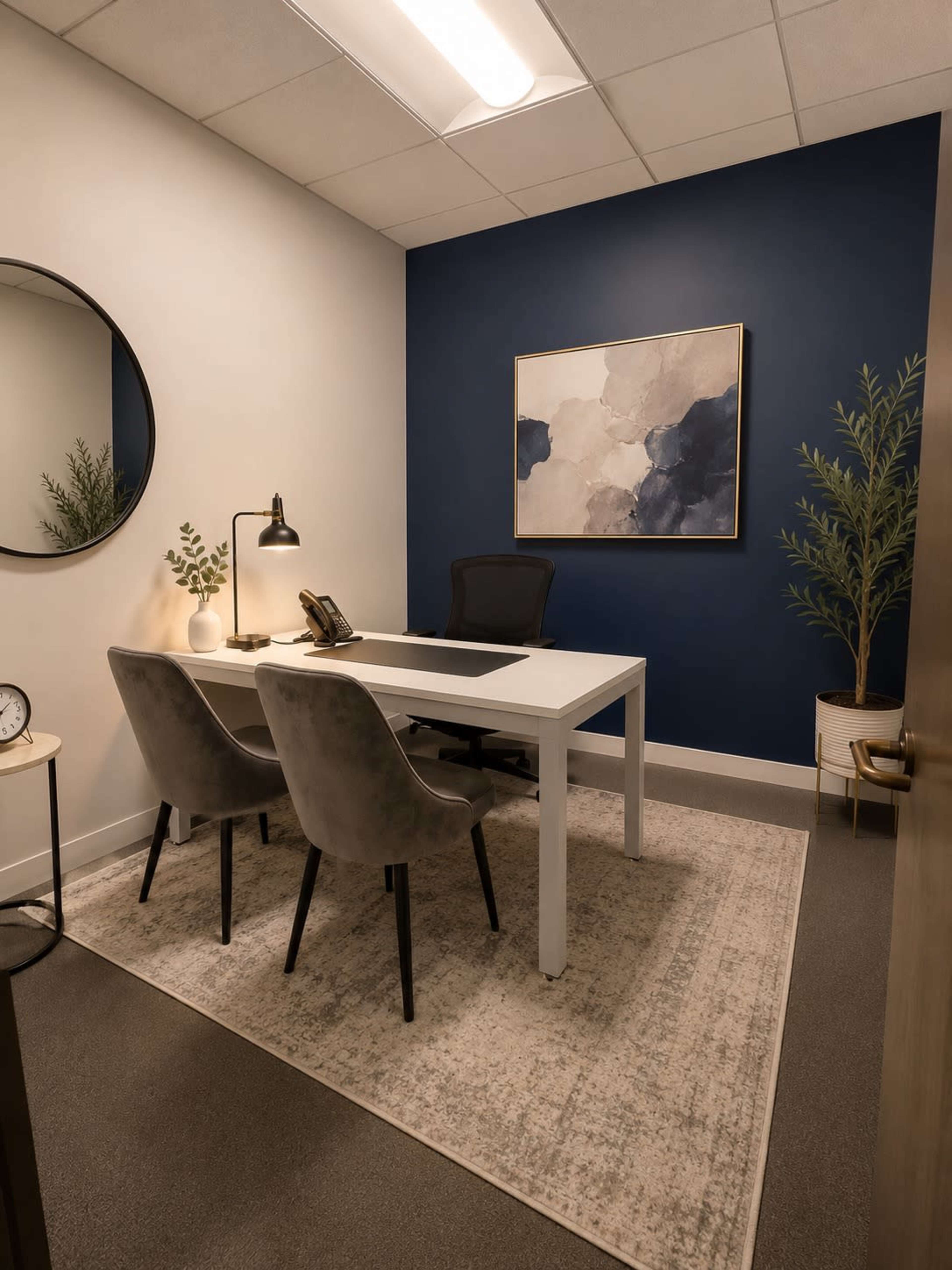 A modern office space features a white desk with two gray chairs, a round mirror on the wall, and a framed abstract painting against a dark blue wall.