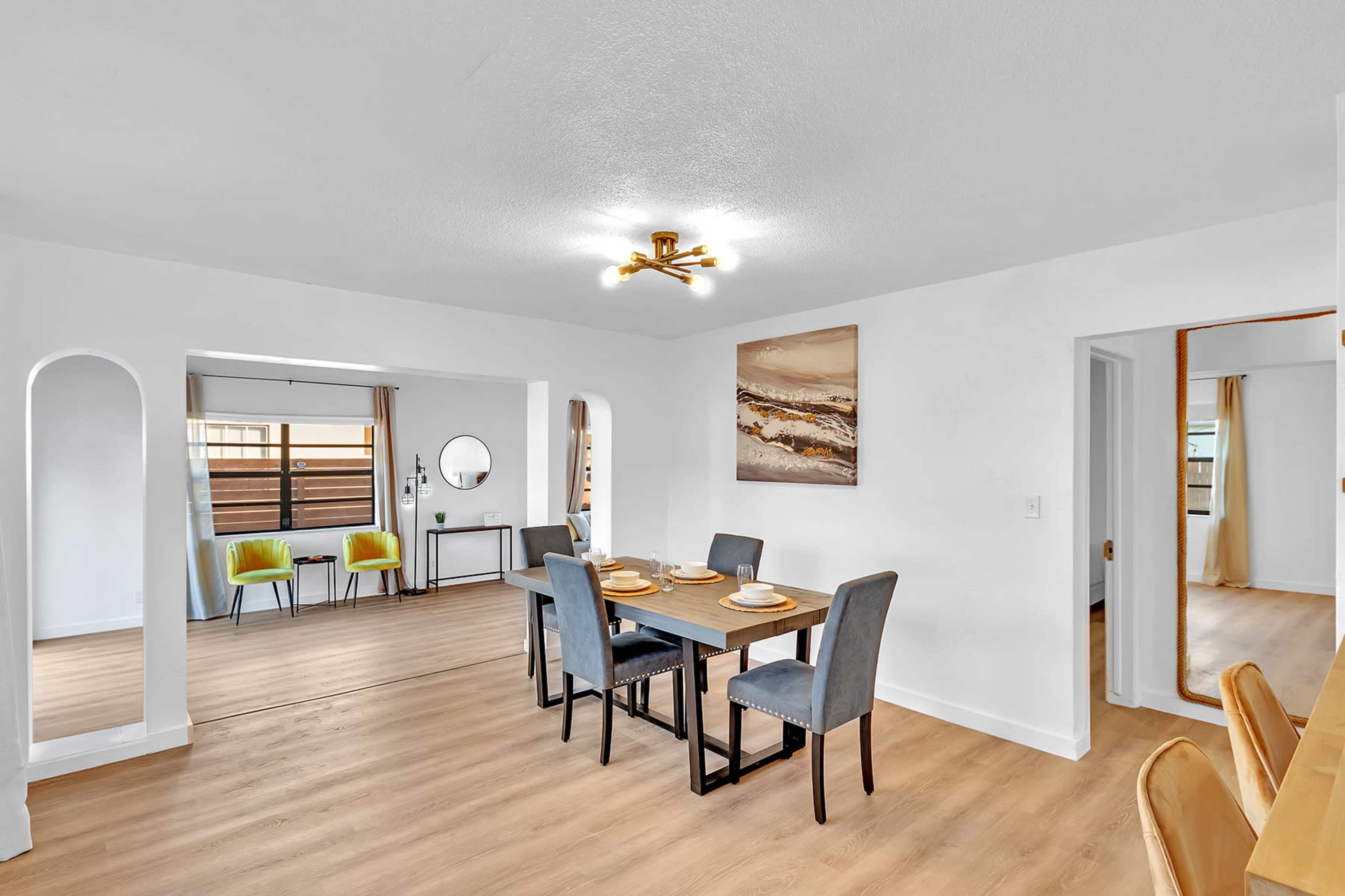 A dining area features a rectangular table set for four, surrounded by upholstered chairs, with a brightly lit open-plan layout leading to a living space.