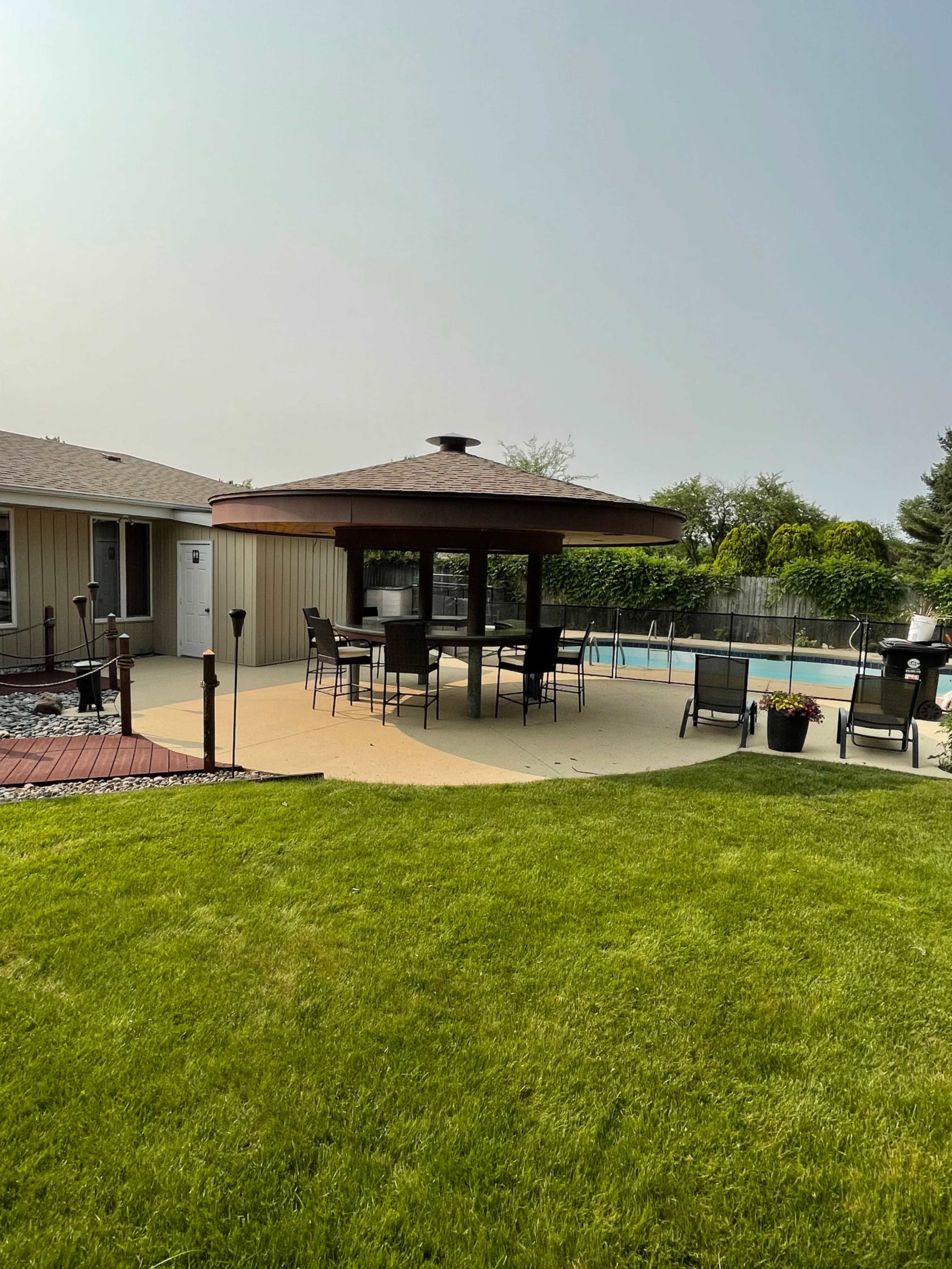 A backyard with a gazebo containing a dining set, surrounding a swimming pool and grassy area.