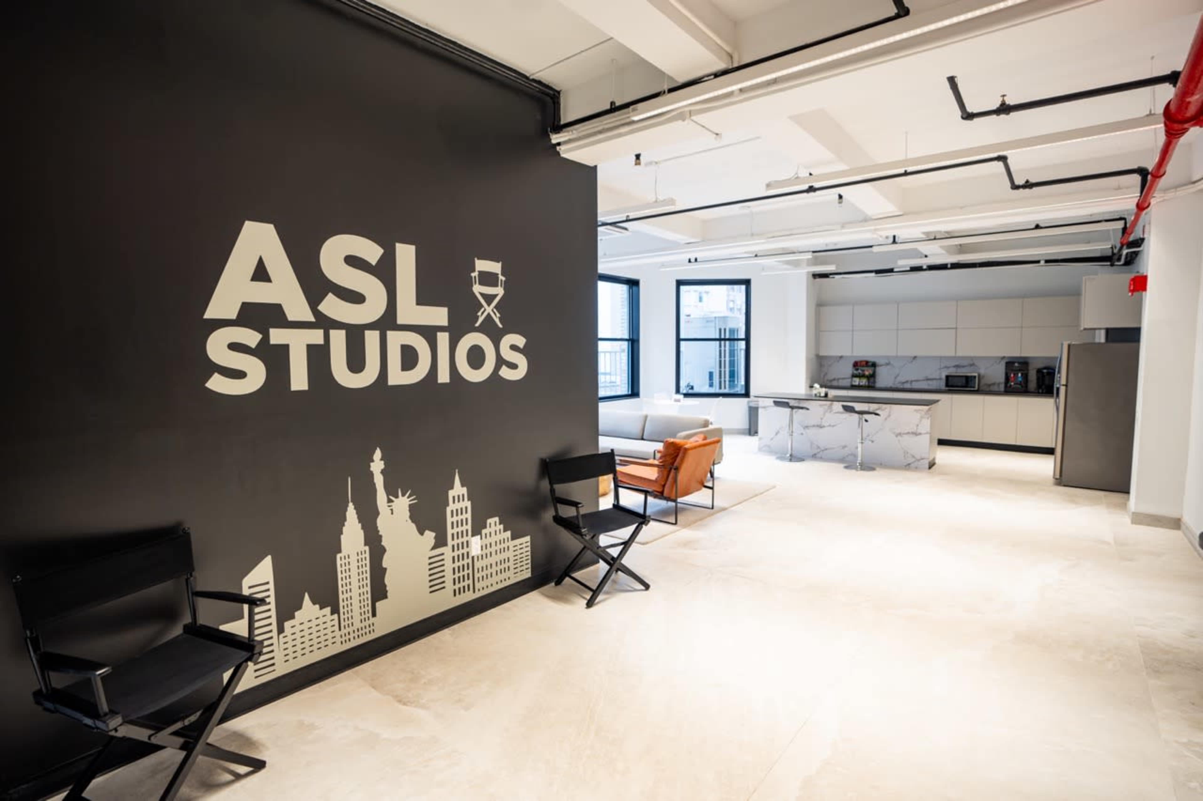 A modern studio space features a dark wall with the text "ASL Studios" and a skyline graphic, alongside a kitchen area and seating.