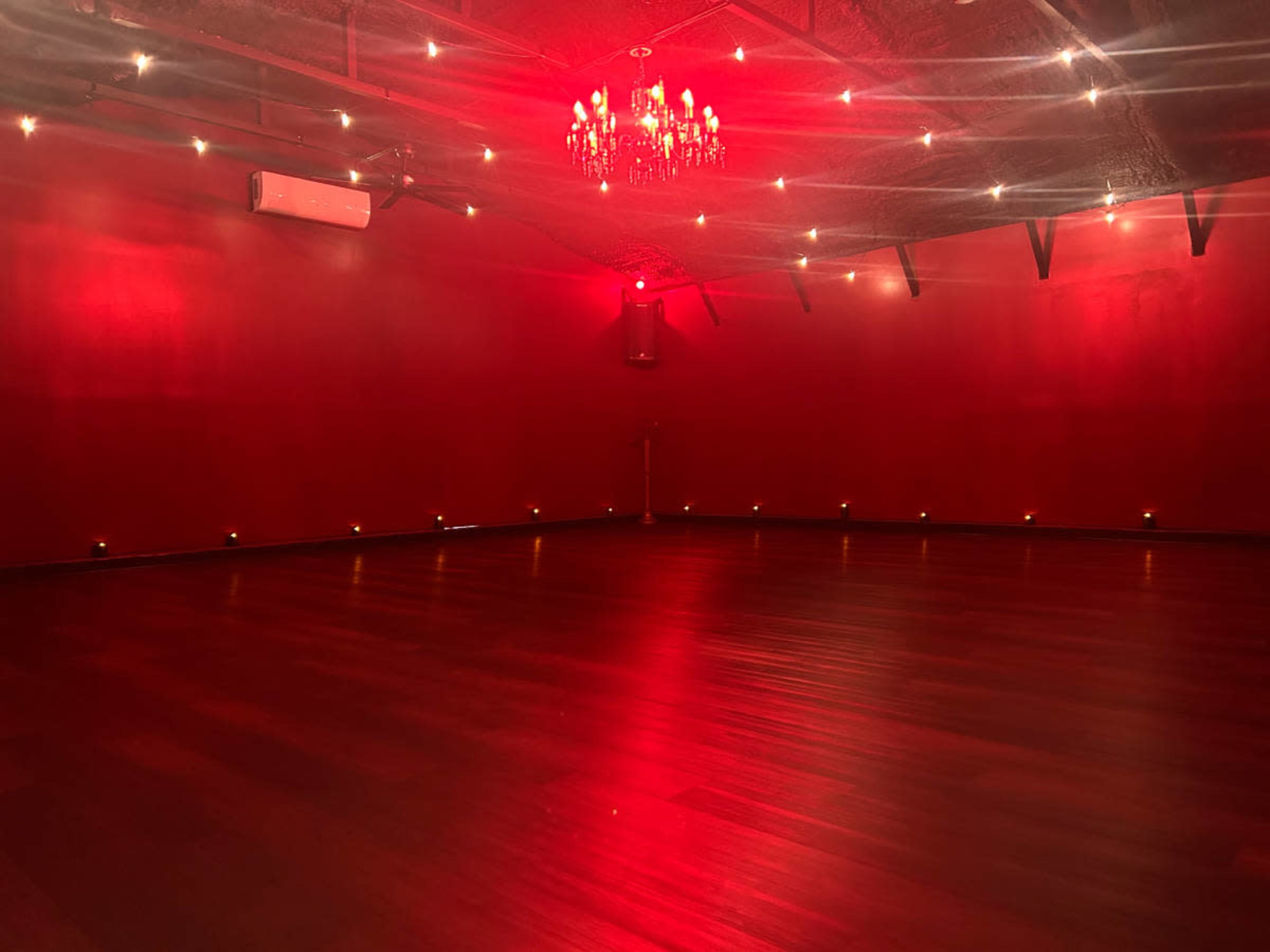 The image shows a dimly lit room with red walls, wooden flooring, and decorative lighting, including a chandelier and wall-mounted lights.