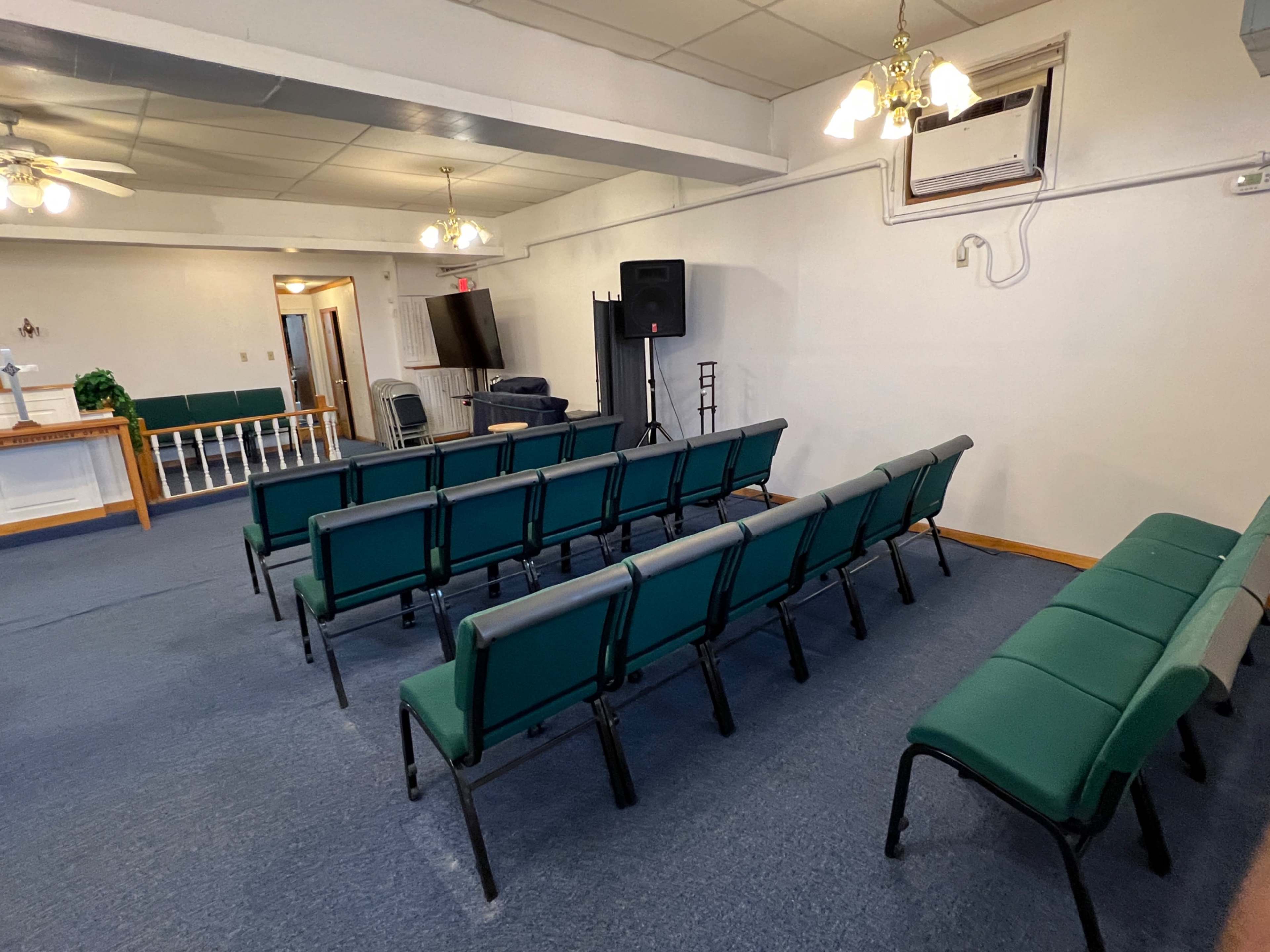 The image shows a sparsely furnished meeting room with rows of green chairs, a small stage area, and a television on one side.