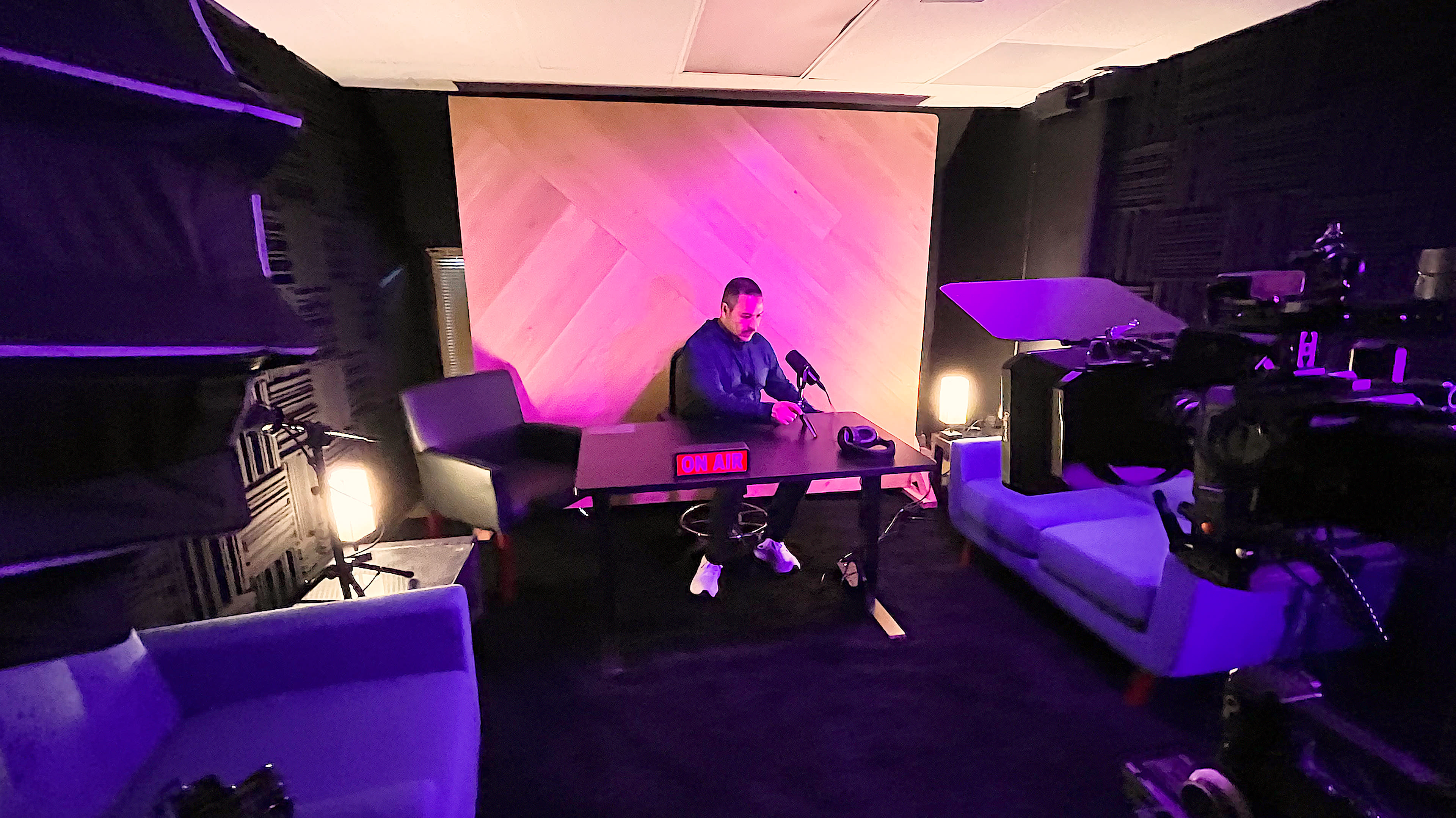 A person sits at a desk in a dimly lit studio with purple lighting, surrounded by two blue chairs and filming equipment.