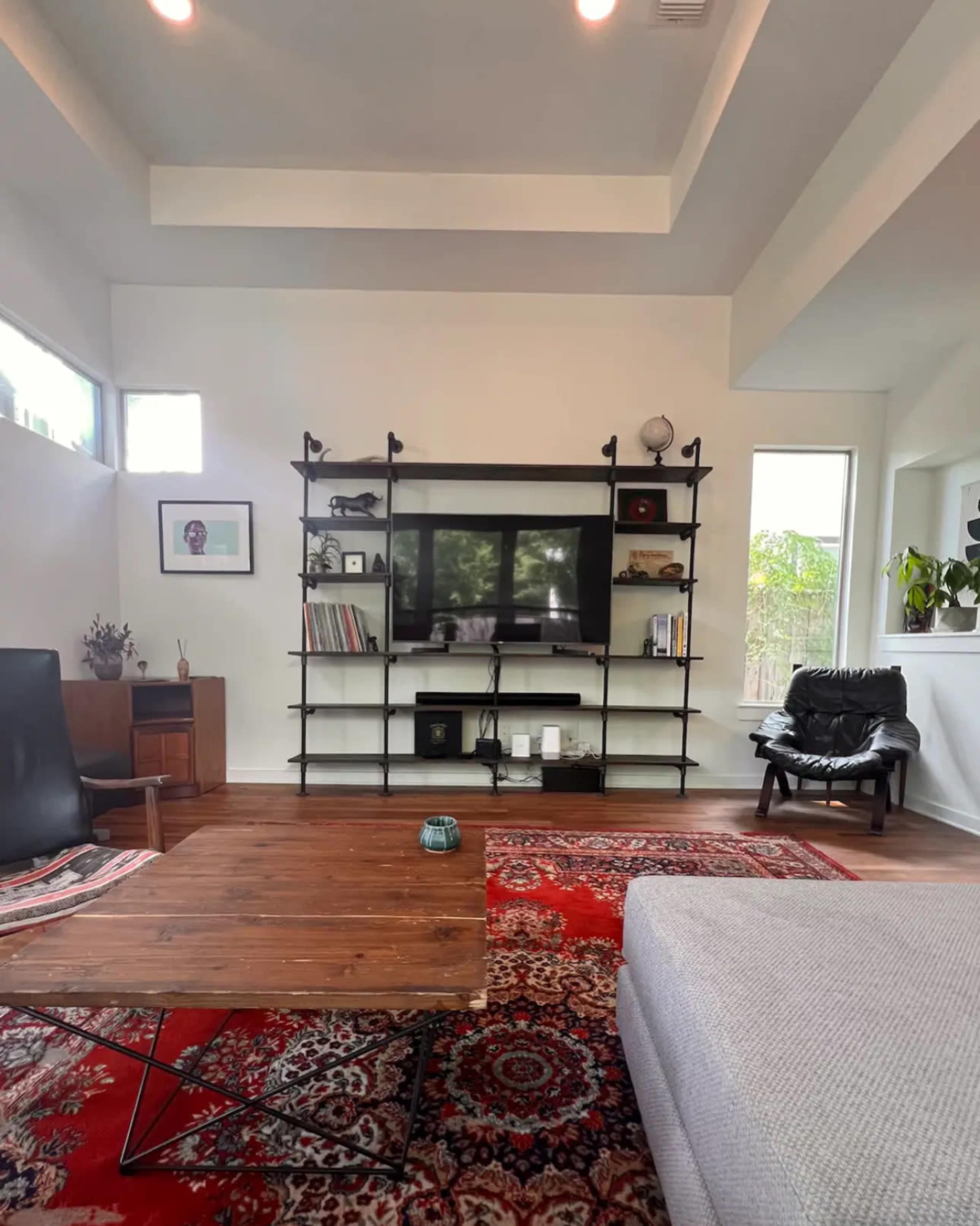 A modern living room with a black metal shelving unit holding a television and books, set against a wall with windows and complemented by a wooden coffee table and a patterned rug.