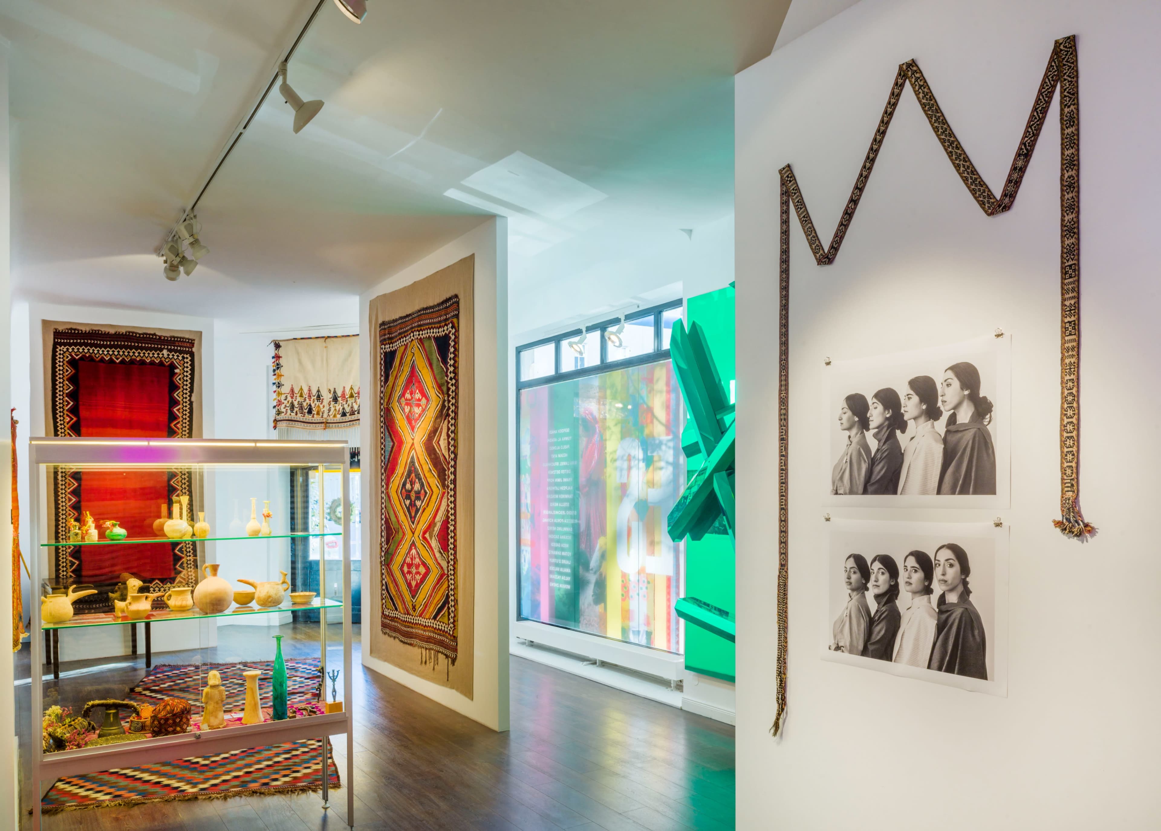 The image shows an exhibition space featuring traditional textiles and artifacts displayed on shelves alongside vibrant wall decorations and photographs of women.