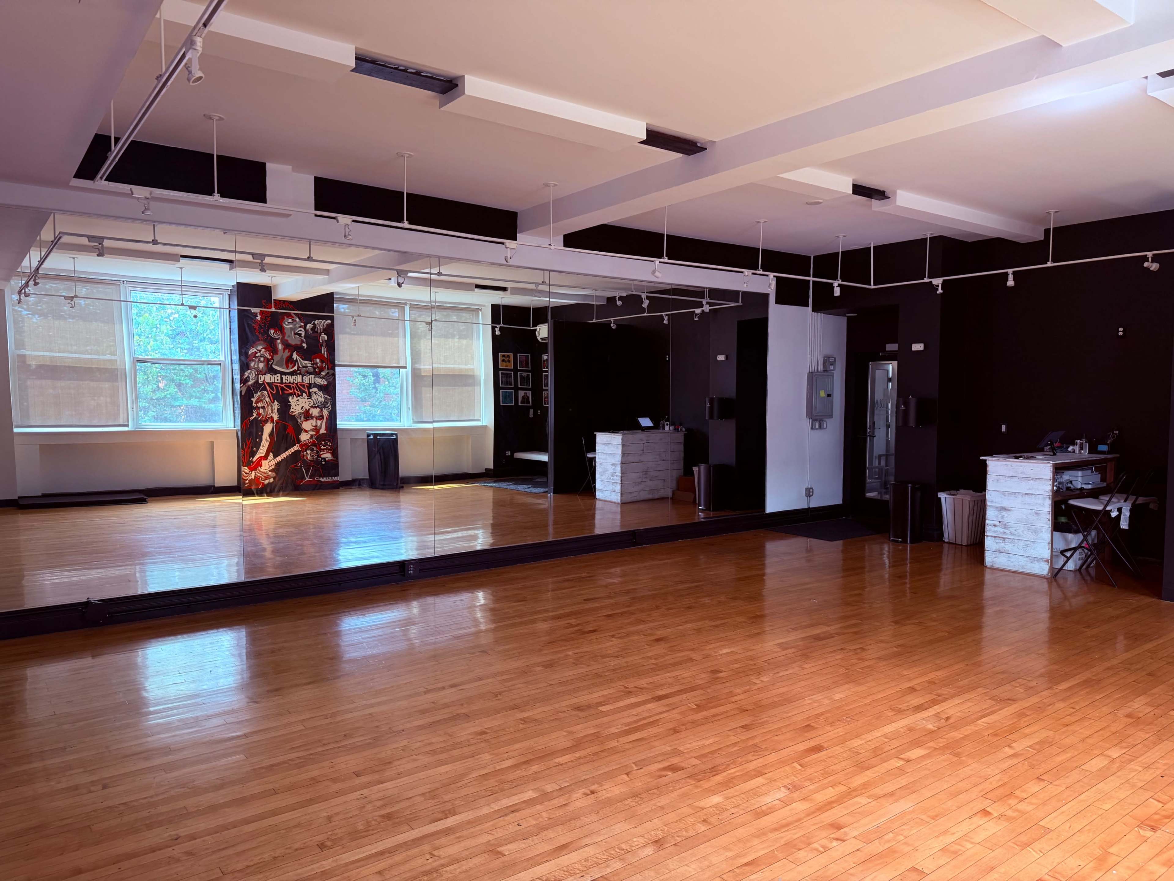 The image shows an empty dance studio with hardwood floors, a large mirror, and windows allowing natural light in, along with wall art in the background.