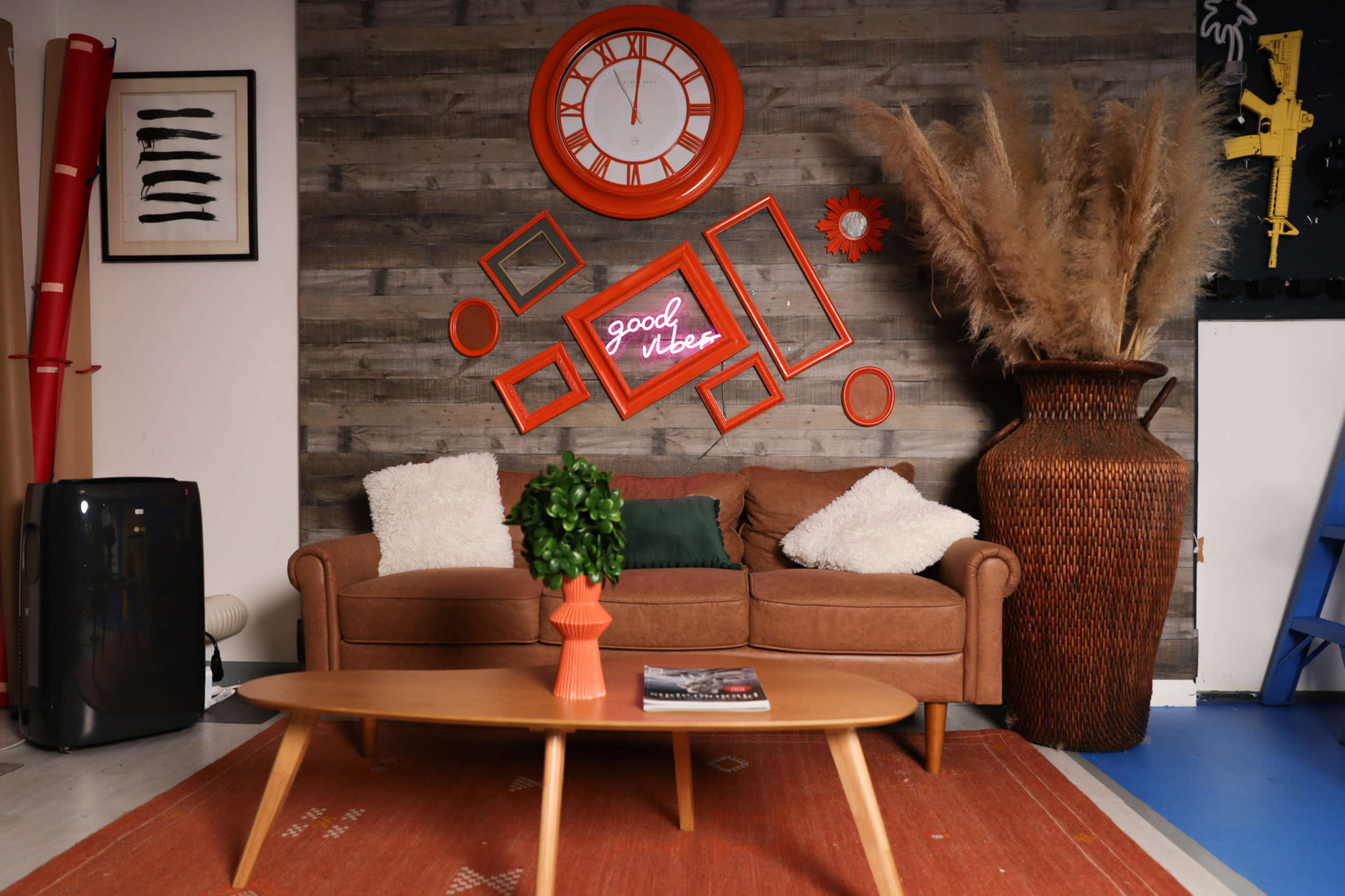The image features a cozy living room scene with a brown sofa adorned with white and green cushions, a wooden coffee table, and an orange wall clock alongside a neon sign saying "good vibes."