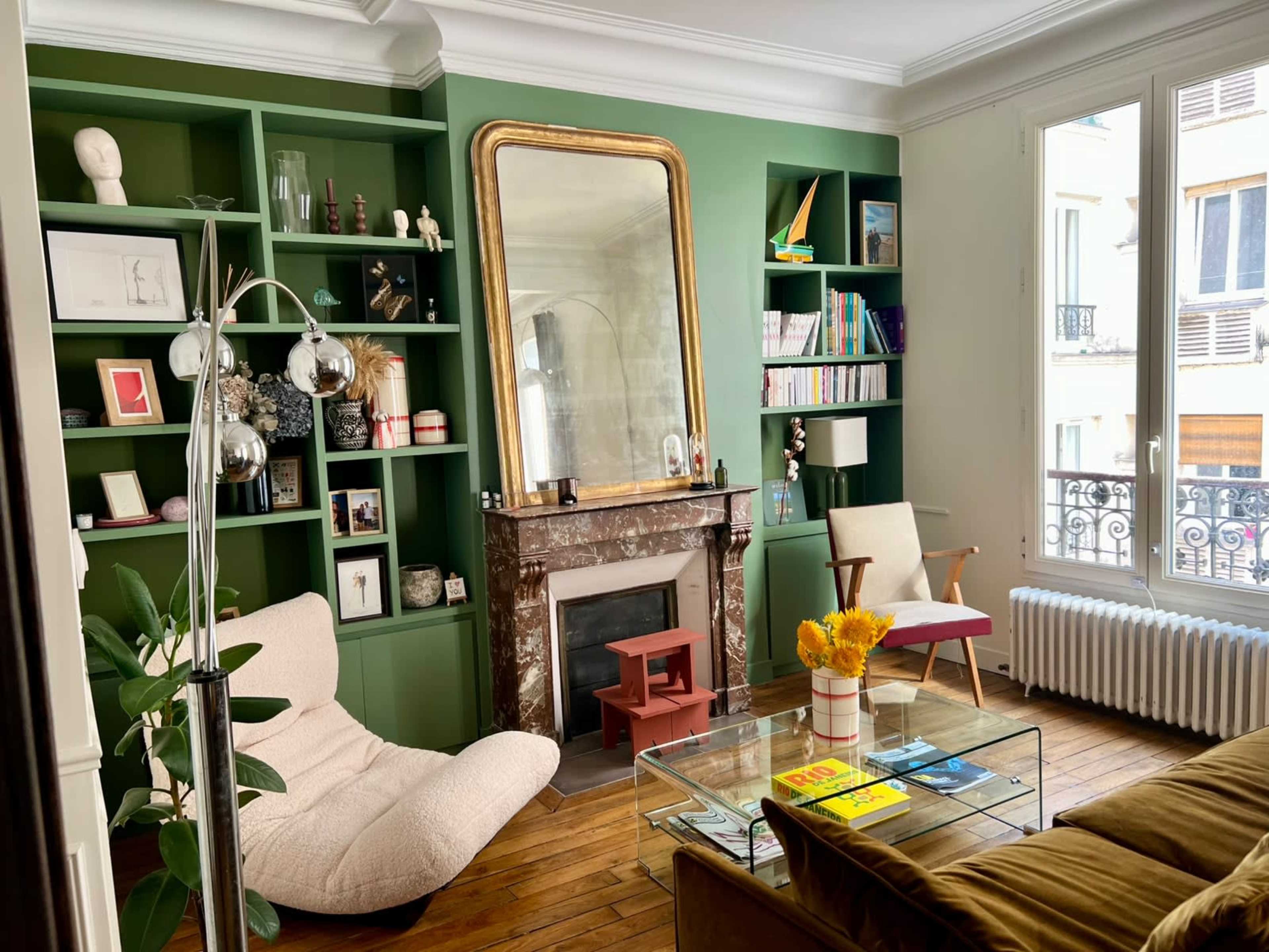 The image shows a stylish living room with green shelving, a large mirror above a marble fireplace, and a cozy seating area that includes a modern chair and a lounge chair.