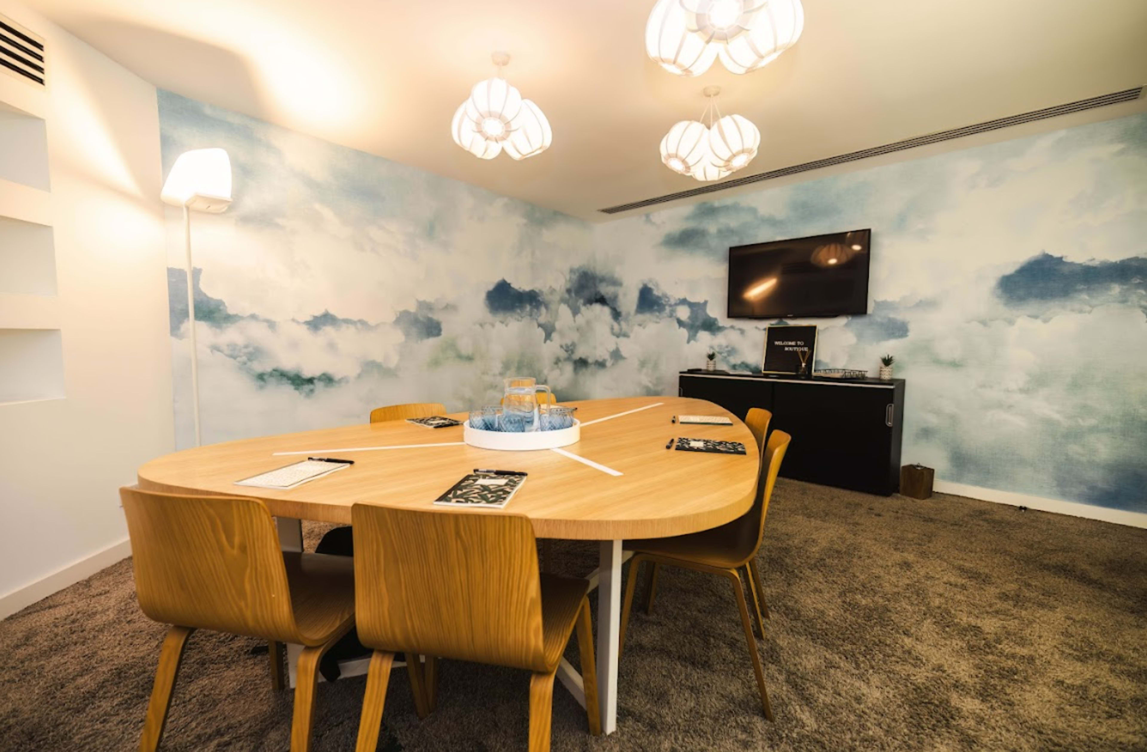 A meeting room features an oval wooden table surrounded by chairs, with a light blue cloud-patterned wall and a television mounted on the opposite wall.