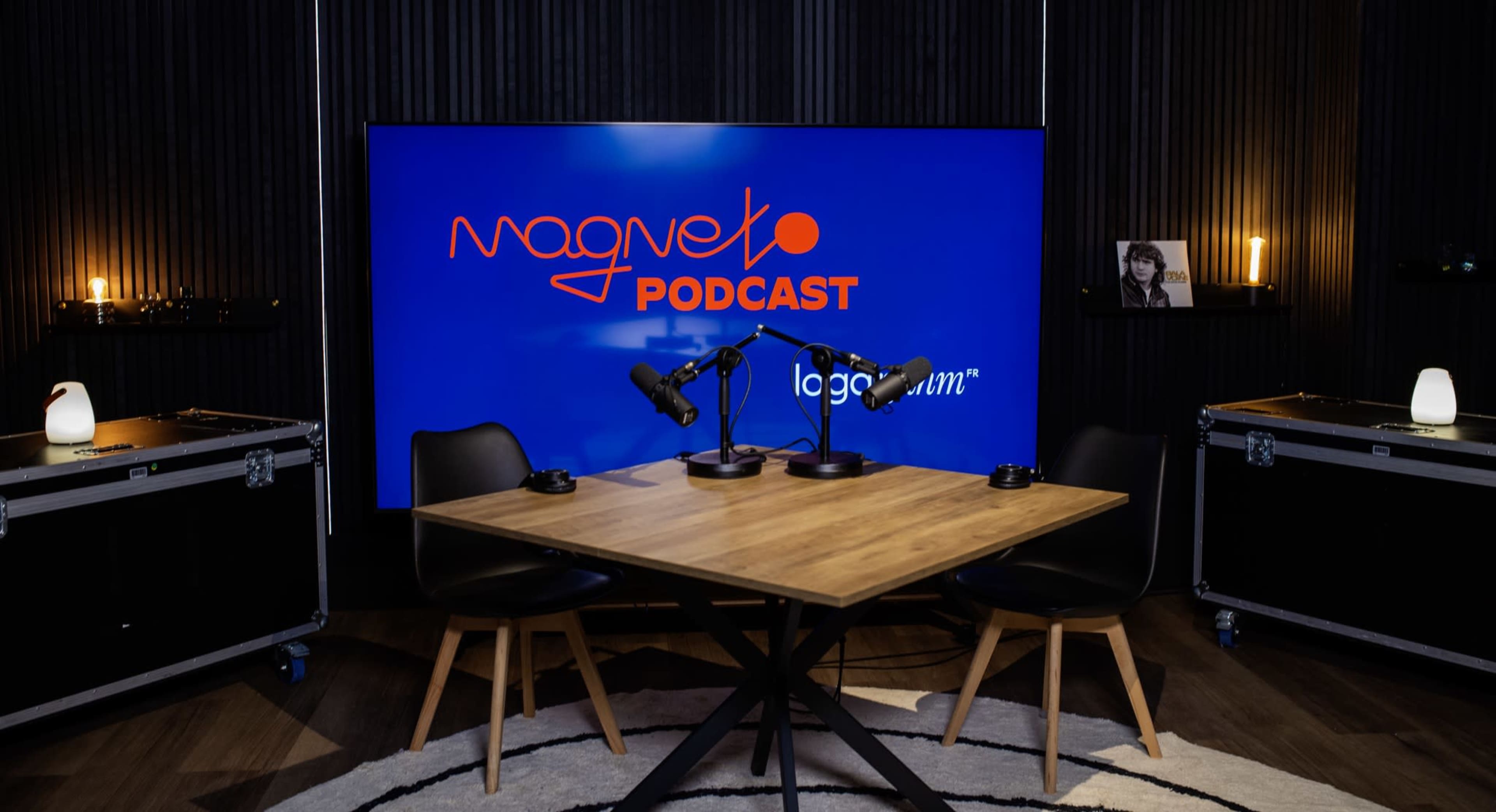 The image shows a podcast recording studio featuring a wooden table with two black chairs, two microphones on boom arms, and a large screen displaying the "Magneto Podcast" logo in a dimly lit environment.