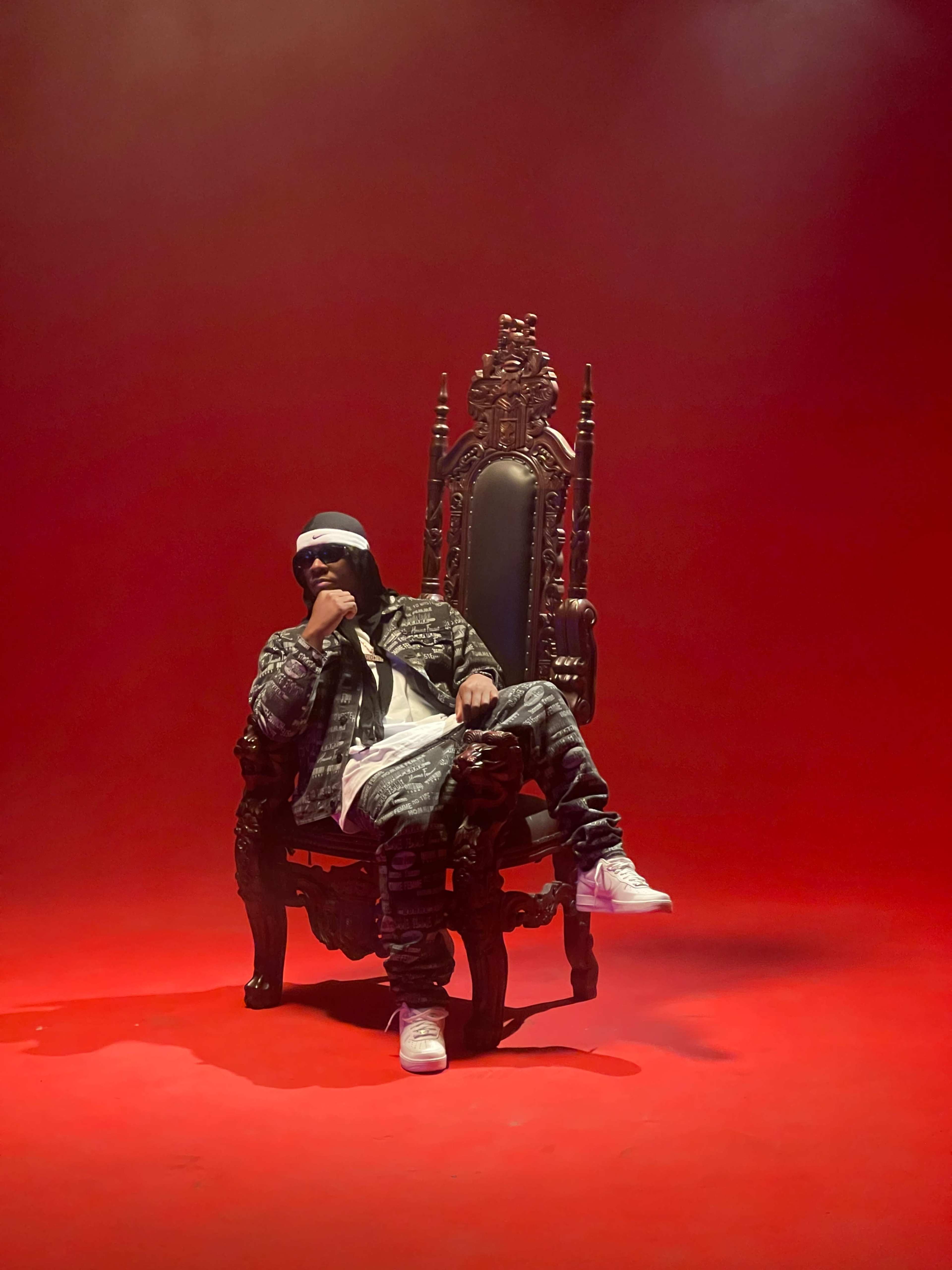 A person wearing a black outfit and white sneakers sits thoughtfully in an ornate throne against a red backdrop.