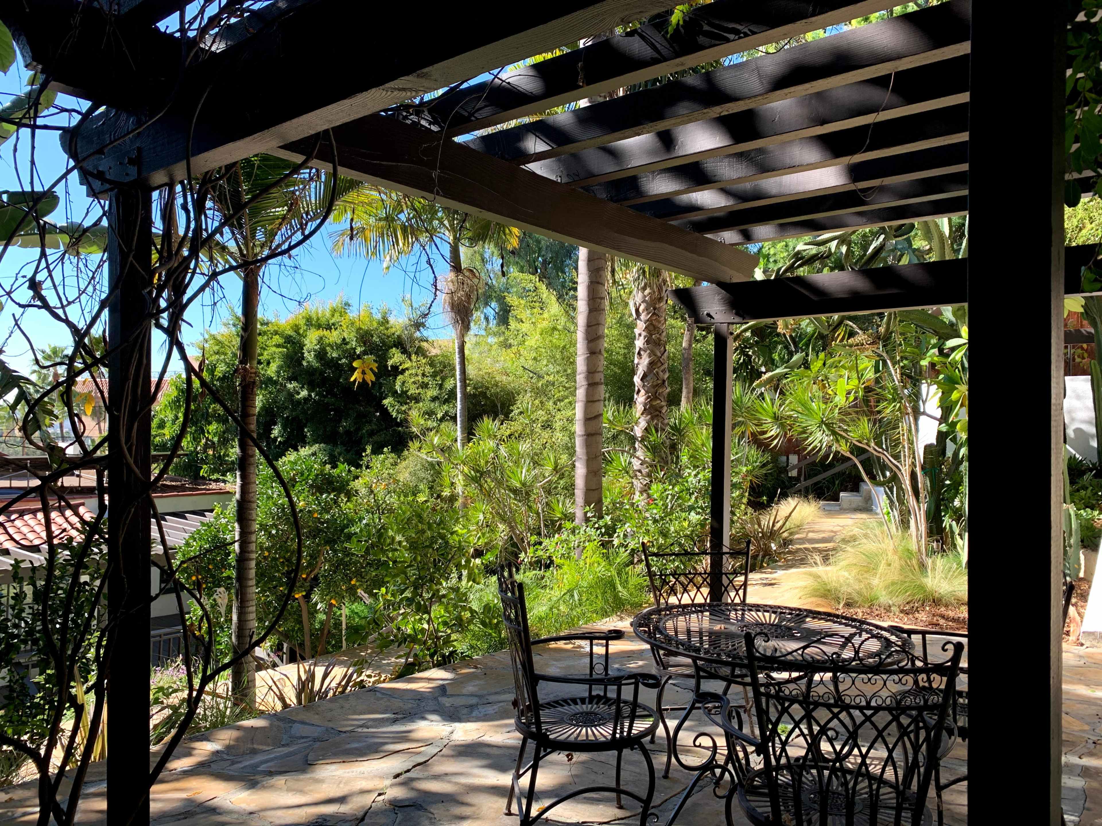 A shaded patio area with wrought iron furniture overlooks a lush garden surrounded by palm trees and greenery.