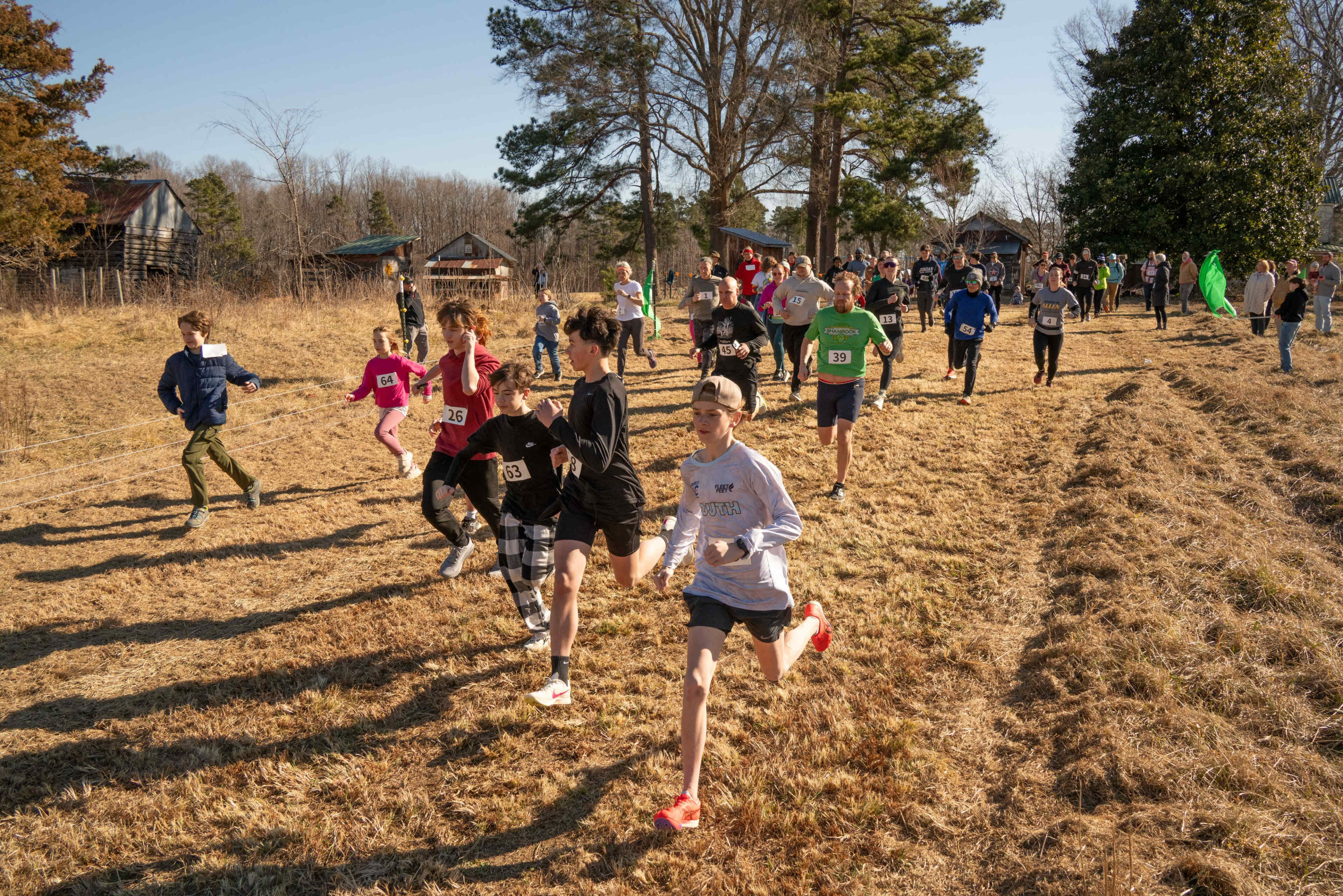 A group of runners in athletic clothing races across a grassy field during an outdoor event.