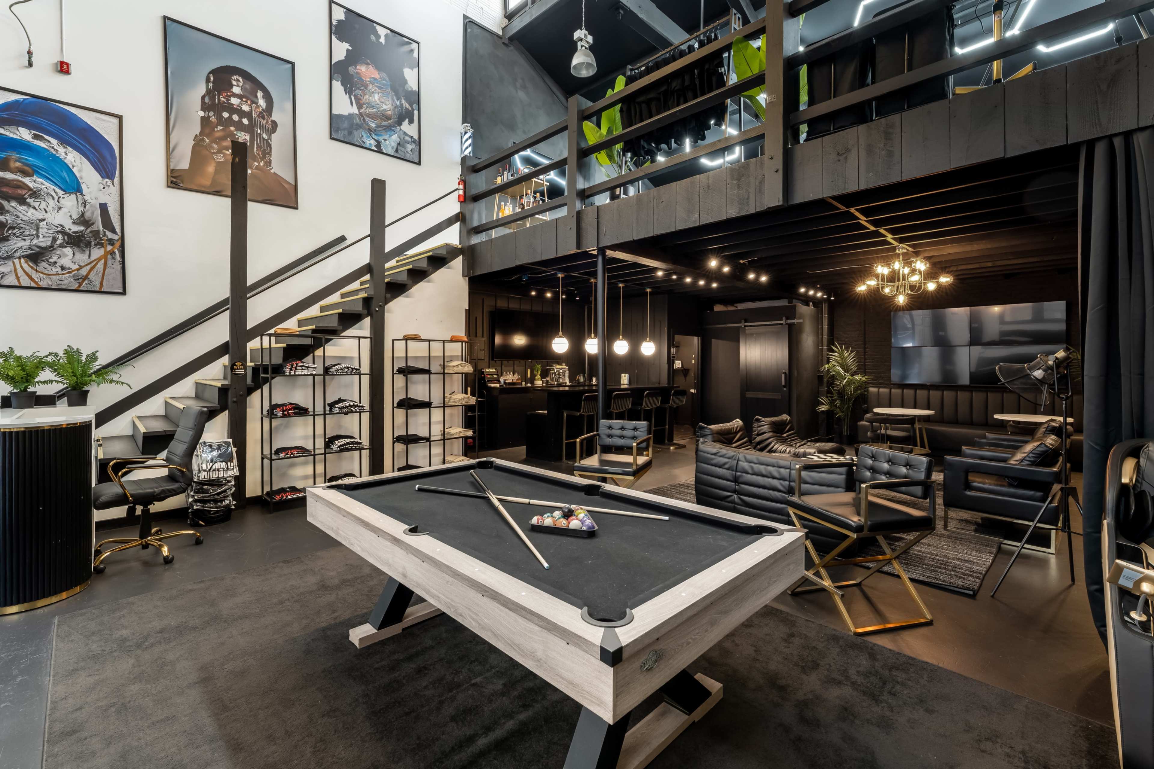 The image shows a modern interior space featuring a billiard table at the center, with a staircase, a lounge area, and various artworks on the walls.