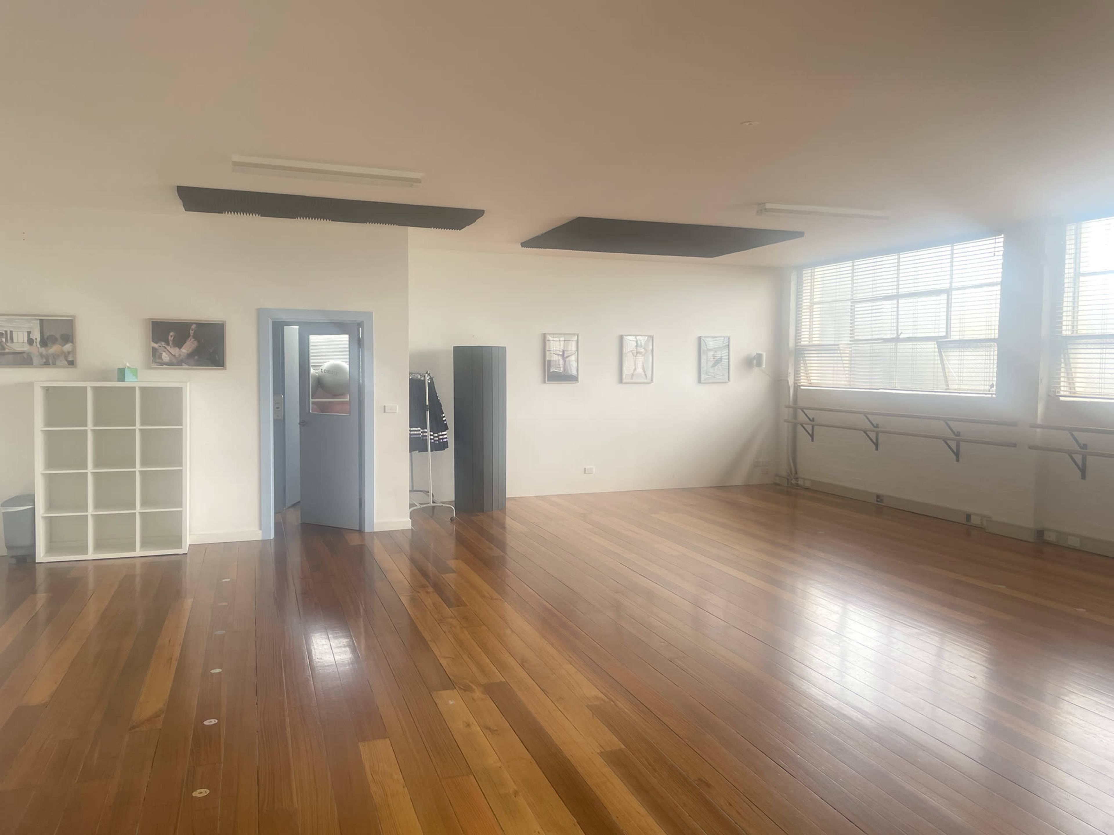 The image shows a spacious studio with polished wooden floors, large windows, and ballet barres along one wall, featuring several framed photos on the adjacent walls.